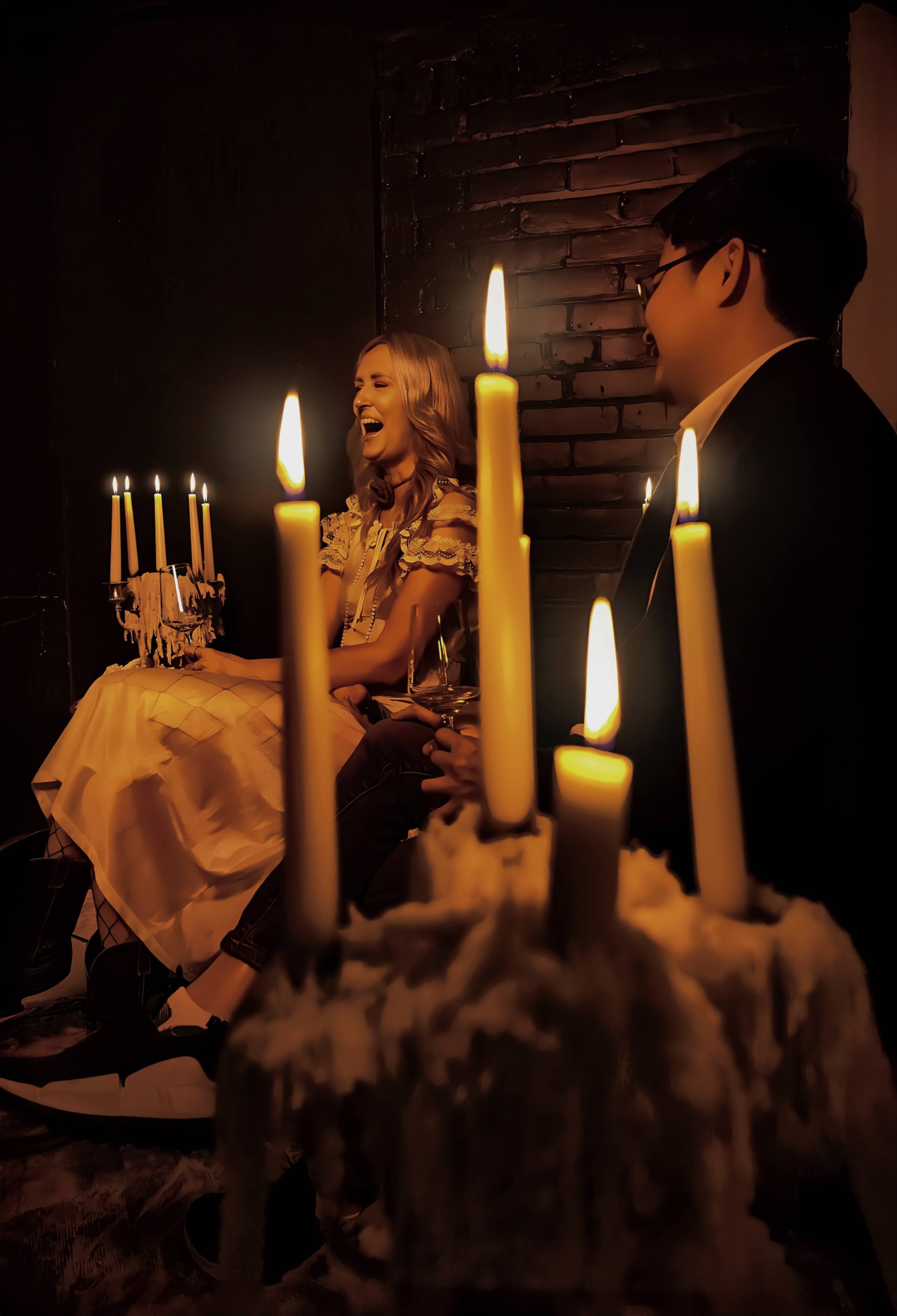 A woman wearing a dress laughs during a birthday celebration with a small chocolate cake with candles and a larger candle-lit candle holder, with a man sitting beside her in a dimly lit room.