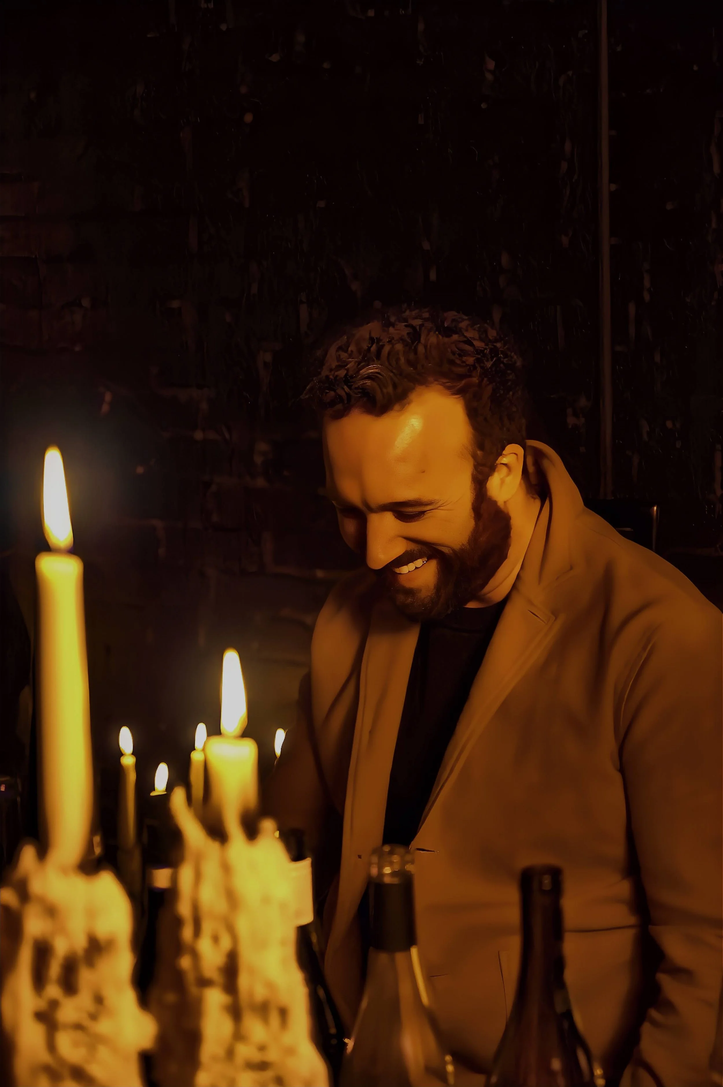 A man with a beard smiling and looking down in a dimly lit room with candles and bottles in front of him.