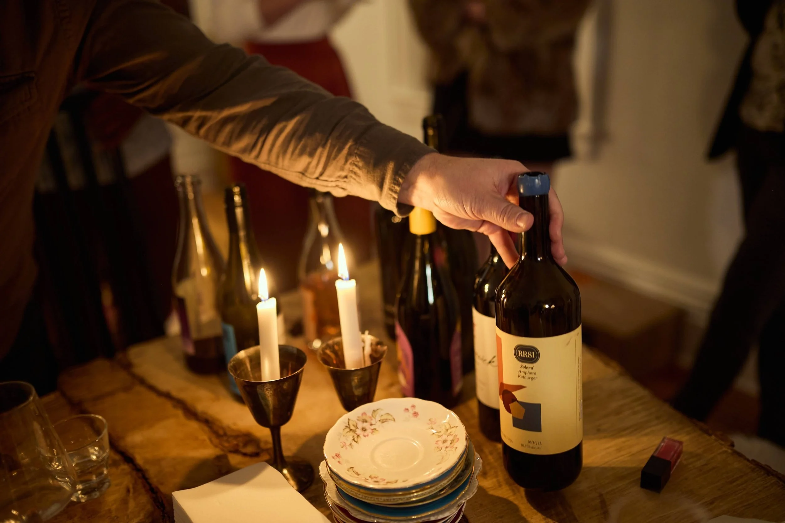 Person reaching for a wine bottle on a table with lit candles, bottles, glasses, and plates, in a dimly lit room.