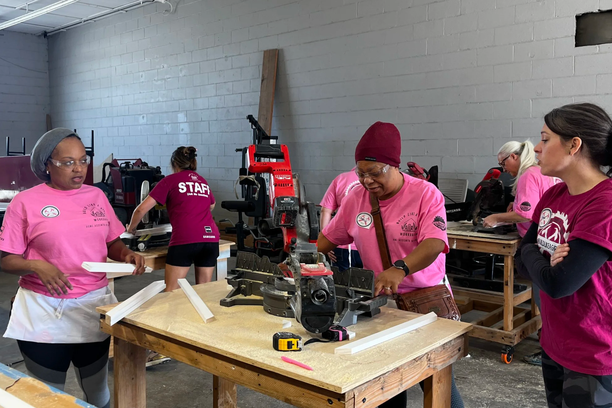 Group of women working on woodworking project in workshop, some cutting and assembling wood, wearing pink shirts and safety glasses.