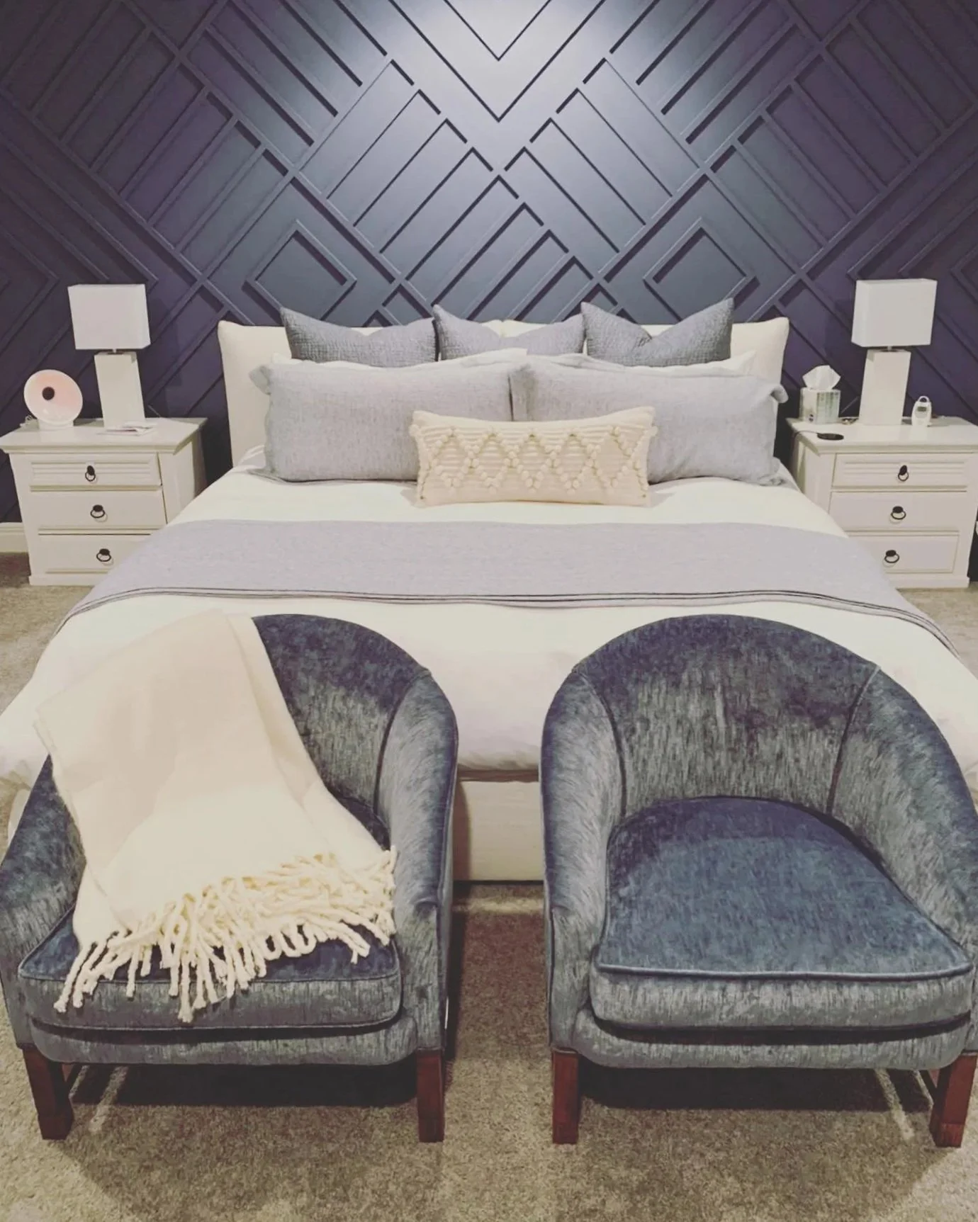 A bedroom with a bed featuring light-colored bedding, gray pillows, and a decorative pillow, flanked by two white nightstands with lamps, against a dark blue, geometric-patterned wall. Two gray velvet chairs are in front of the bed, one with a cream-colored throw blanket.