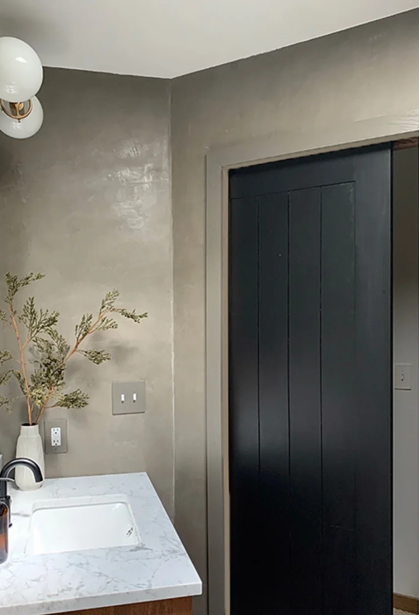 Section of a kitchen with beige textured walls, a white marble countertop, a black door or cabinet, and a vase with dried branches on the counter.
