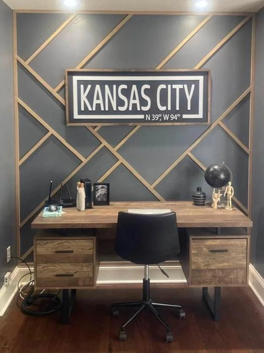 A modern office desk with a black office chair, decorative items, and a framed 'Kansas City' sign on a gray wall with diagonal wood paneling.