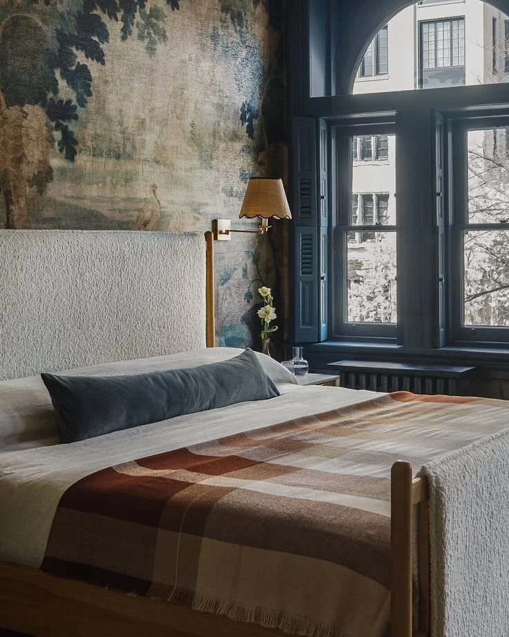 “Vintage-inspired bedroom with mural wall tapestry, cozy plaid wool blanket, textured headboard, and blue arched window shutters in historic townhouse interior”