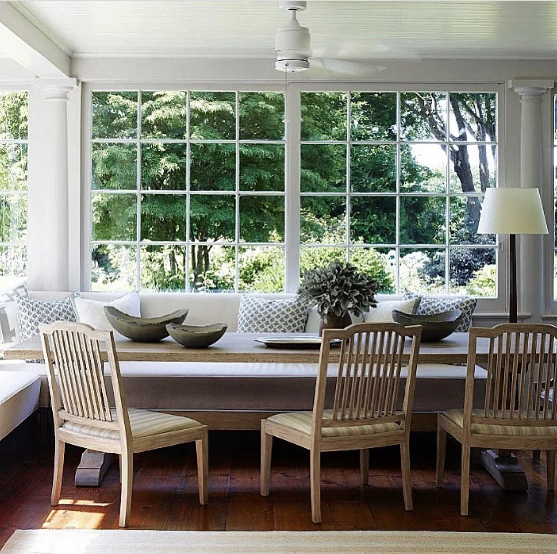 Bright sunroom dining area with wall-to-wall grid windows, built-in bench seating, wood chairs, and views of lush green trees.