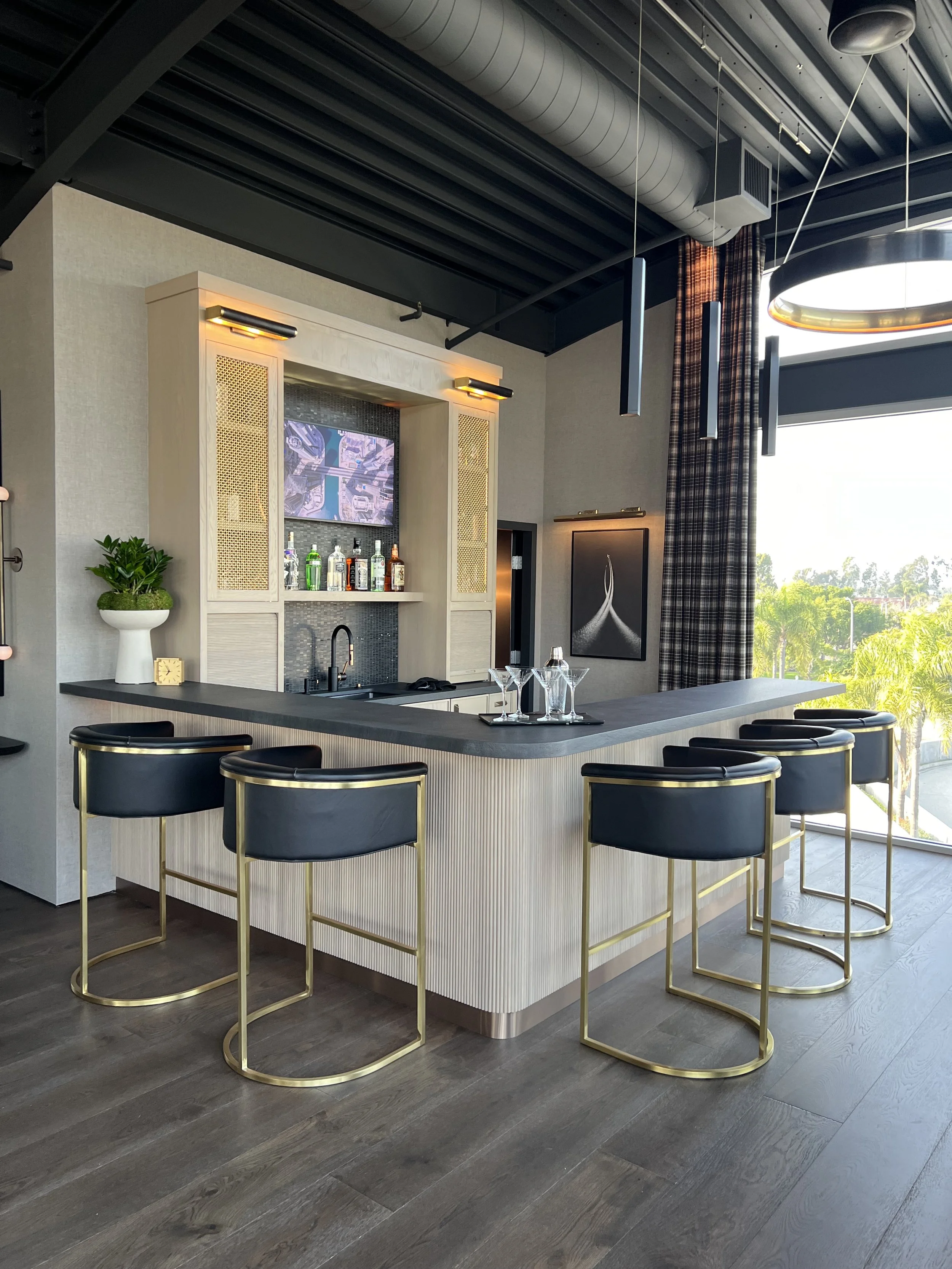 Modern executive bar with gold-accented stools, liquor display, and built-in shelving in daylight