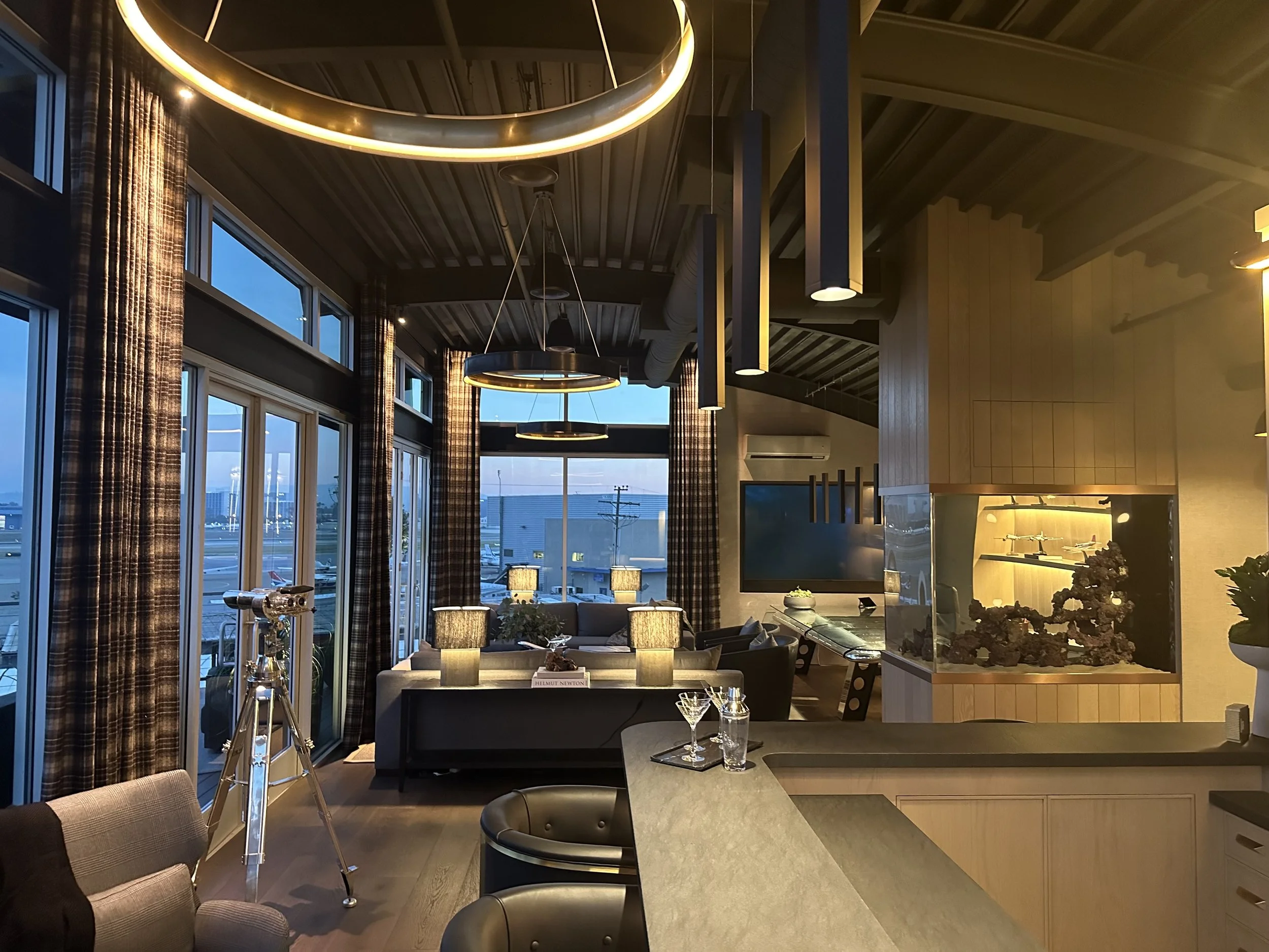 Luxury CEO lounge at dusk with telescope, aquarium, and ambient lighting overlooking private jet runway