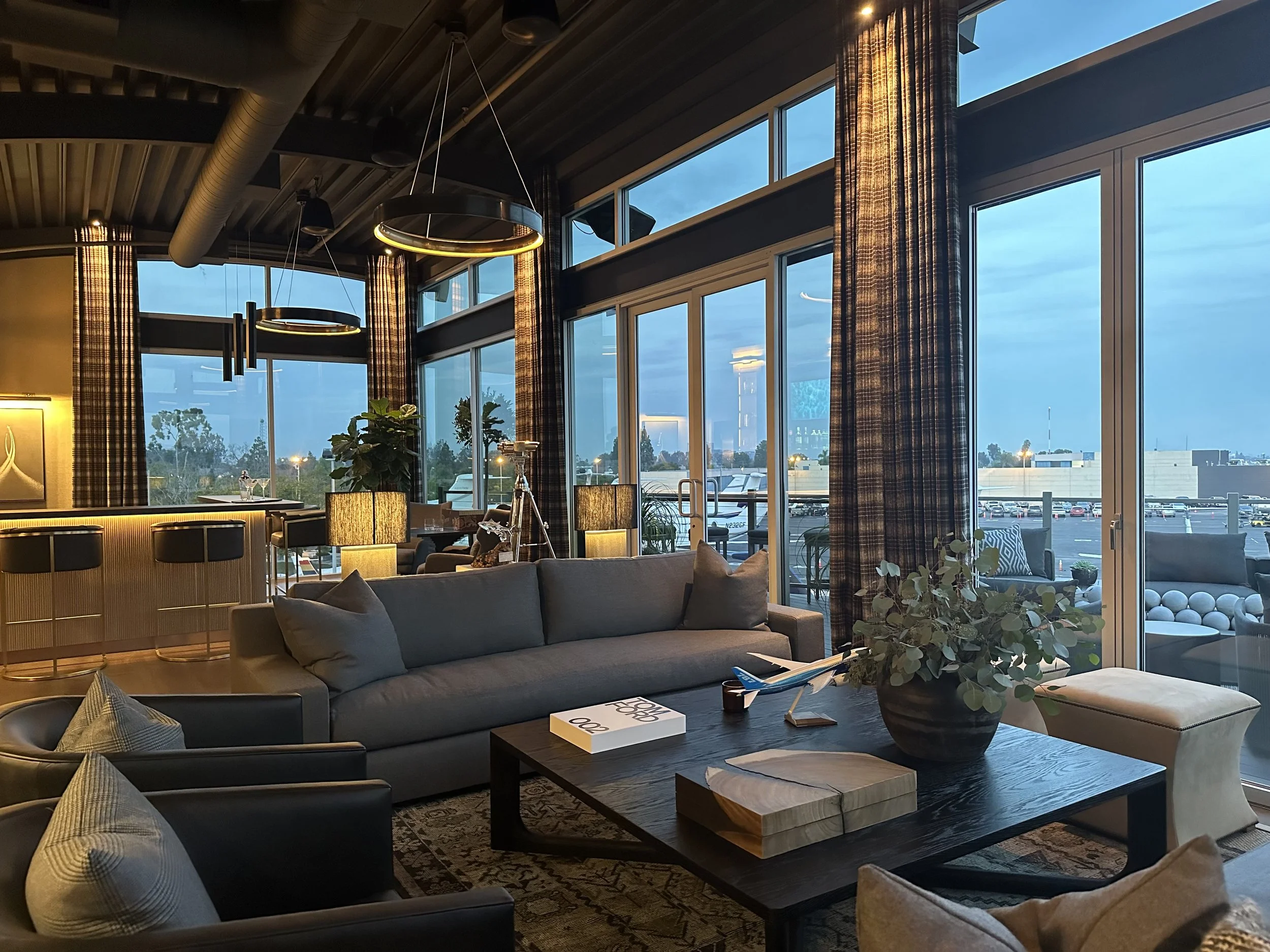 Executive lounge at sunset with grey sofas, stylish lighting, and panoramic windows overlooking private jets