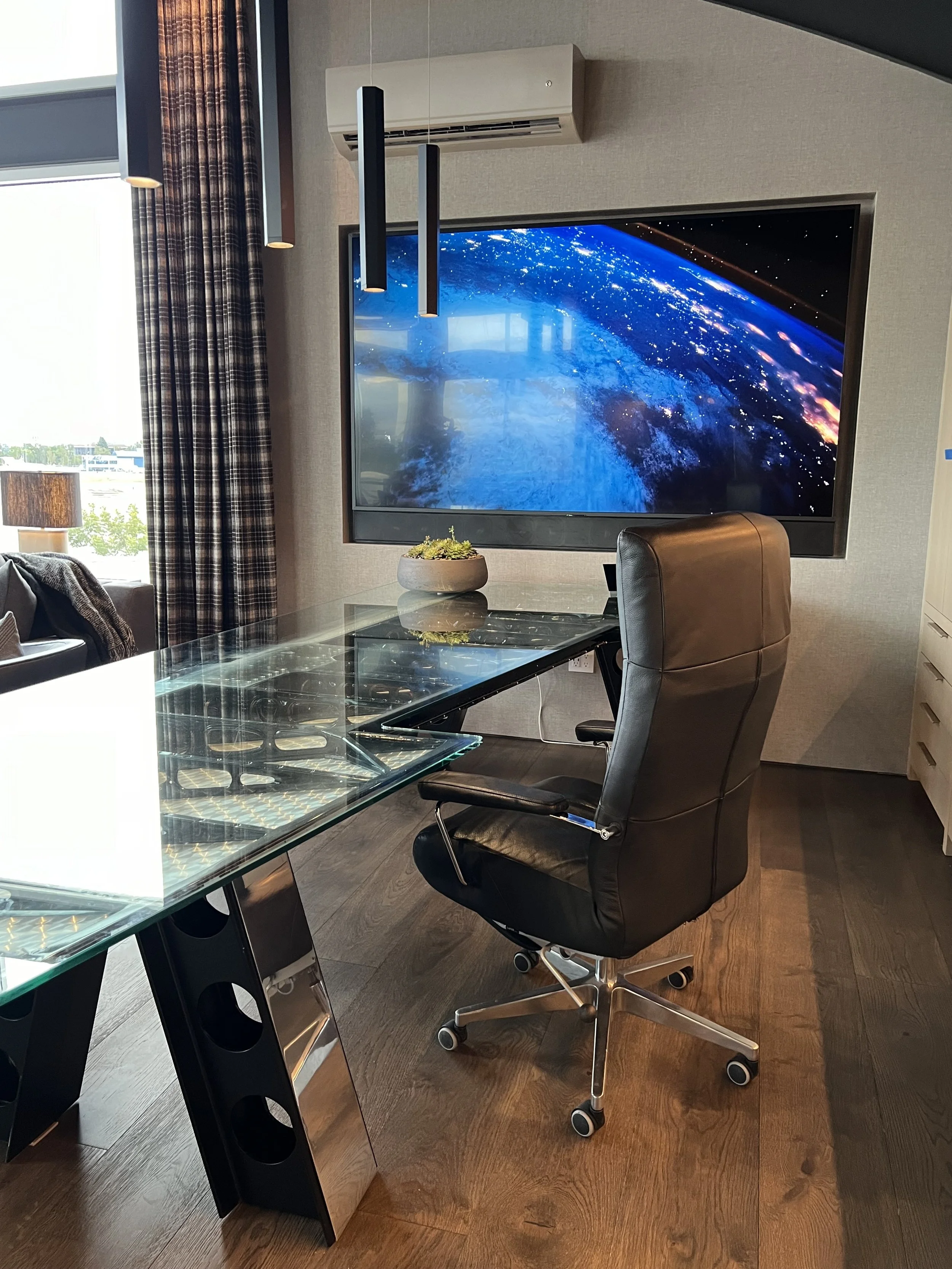 An aviation-wing-inspired desk with a glass top anchors the workspace. A wall-mounted widescreen display and pendant lighting create a perfect blend of functionality and visual impact.