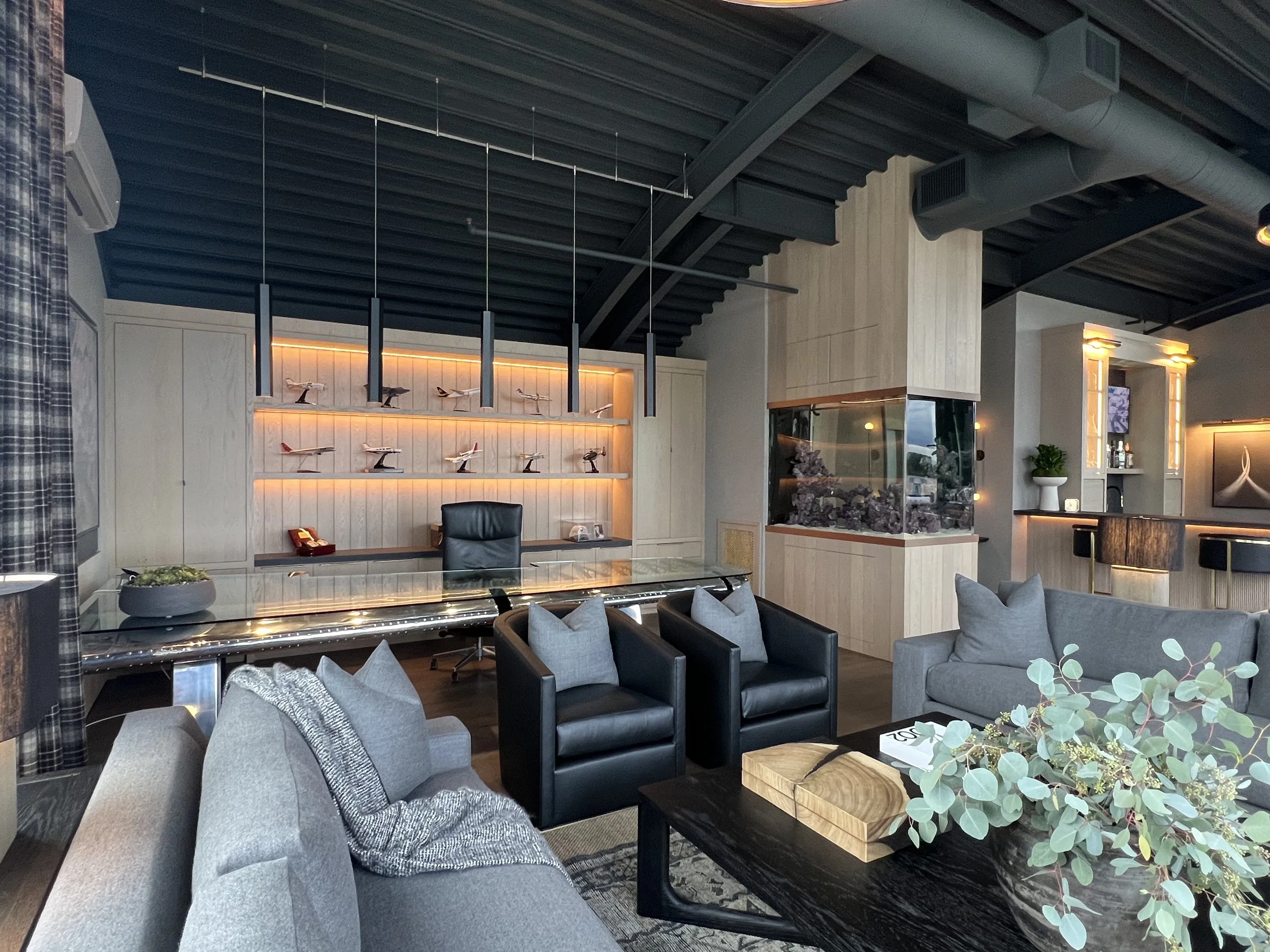 Executive office with modern lounge, large TV, backlit model airplane shelf, and glass executive desk
