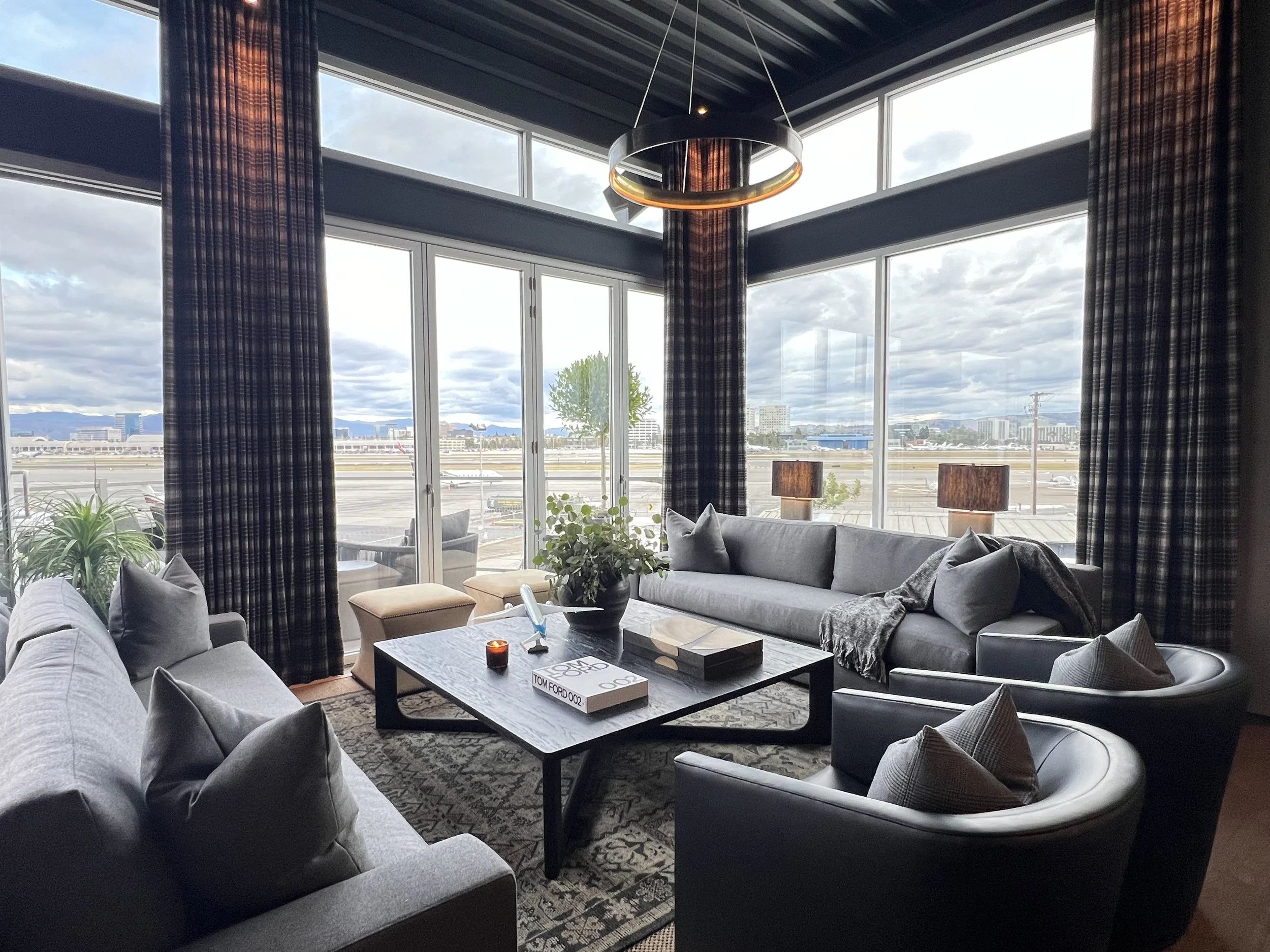 The spacious layout features plush gray sofas, sleek leather chairs, and a black oak coffee table adorned with curated decor, including model aircraft and design books like Tom Ford 002