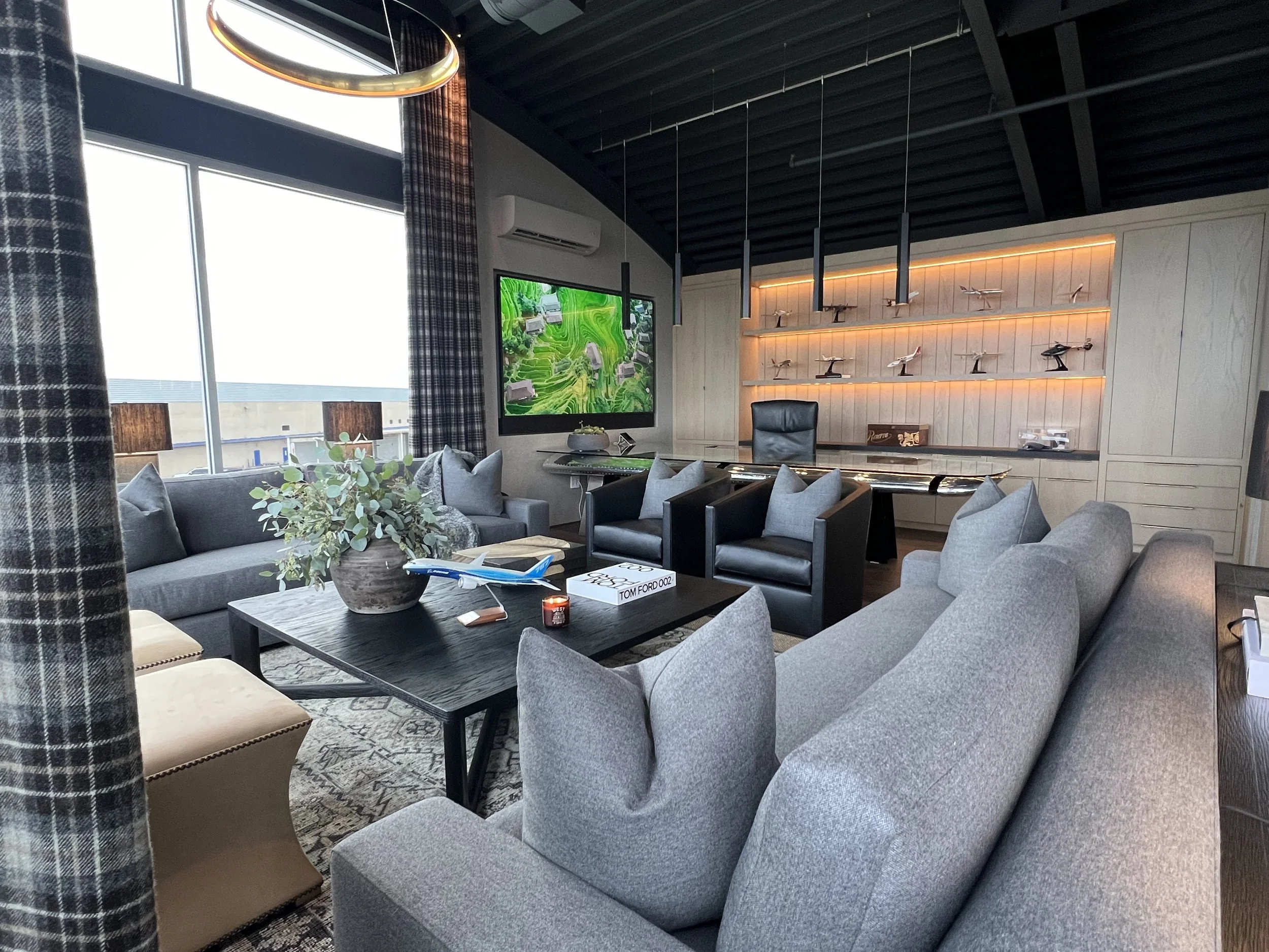 Executive office with modern lounge, large TV, backlit model airplane shelf, and glass executive desk