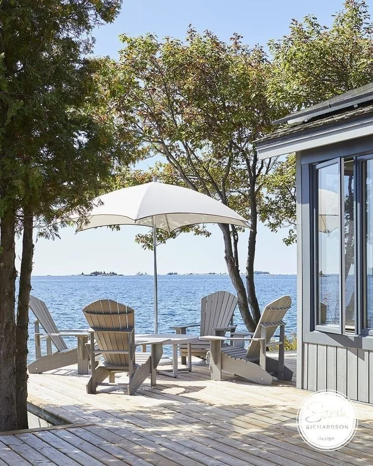 “Tranquil lakeside deck with Adirondack chairs, a white patio umbrella, and surrounding trees overlooking calm blue water.”.jpg