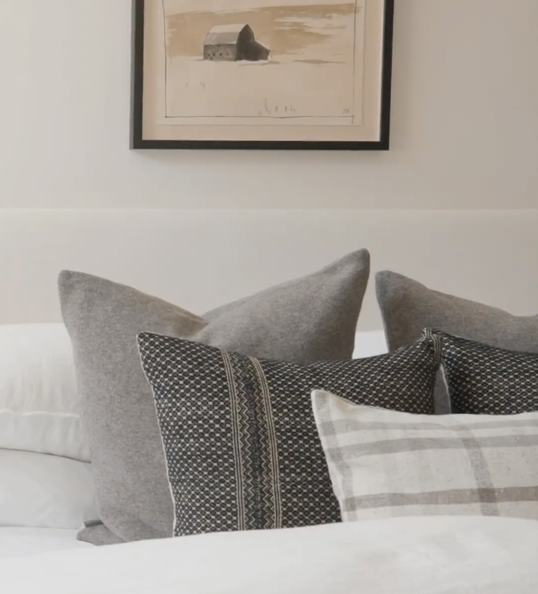 "Modern neutral bedroom with layered throw pillows in grey, black geometric, and plaid patterns beneath rustic barn artwork"