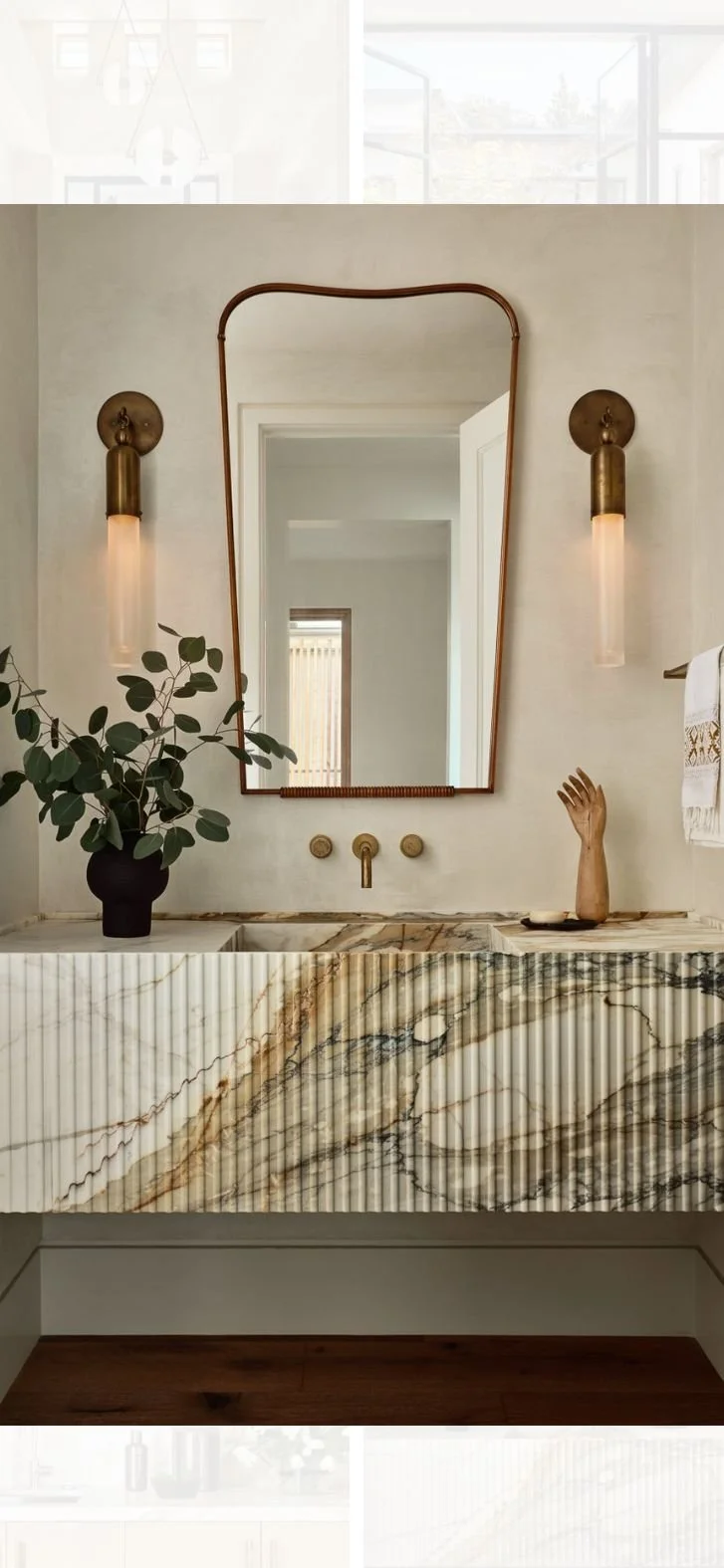Modern powder room with fluted marble vanity, sculptural brass wall sconces, organic-shaped mirror, and minimalist warm-toned palette..jpg