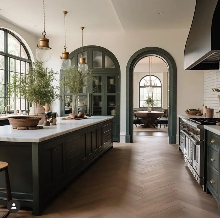 Sophisticated moody kitchen with dark green cabinets, white countertops, arched windows, and pendant lighting