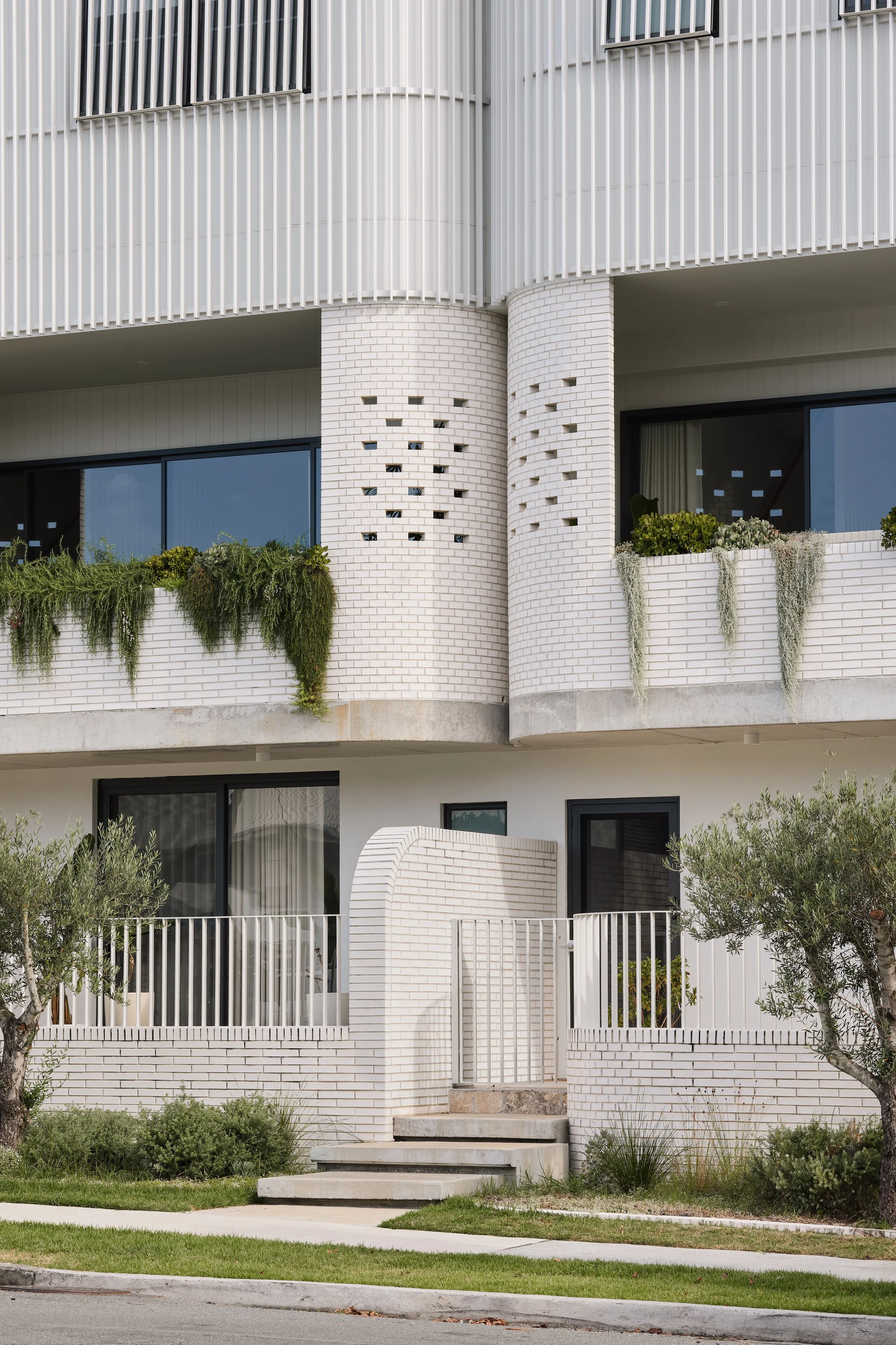 Modern multi-story residential building with white brick exterior, large glass windows, balconies with potted plants, and landscaped greenery.