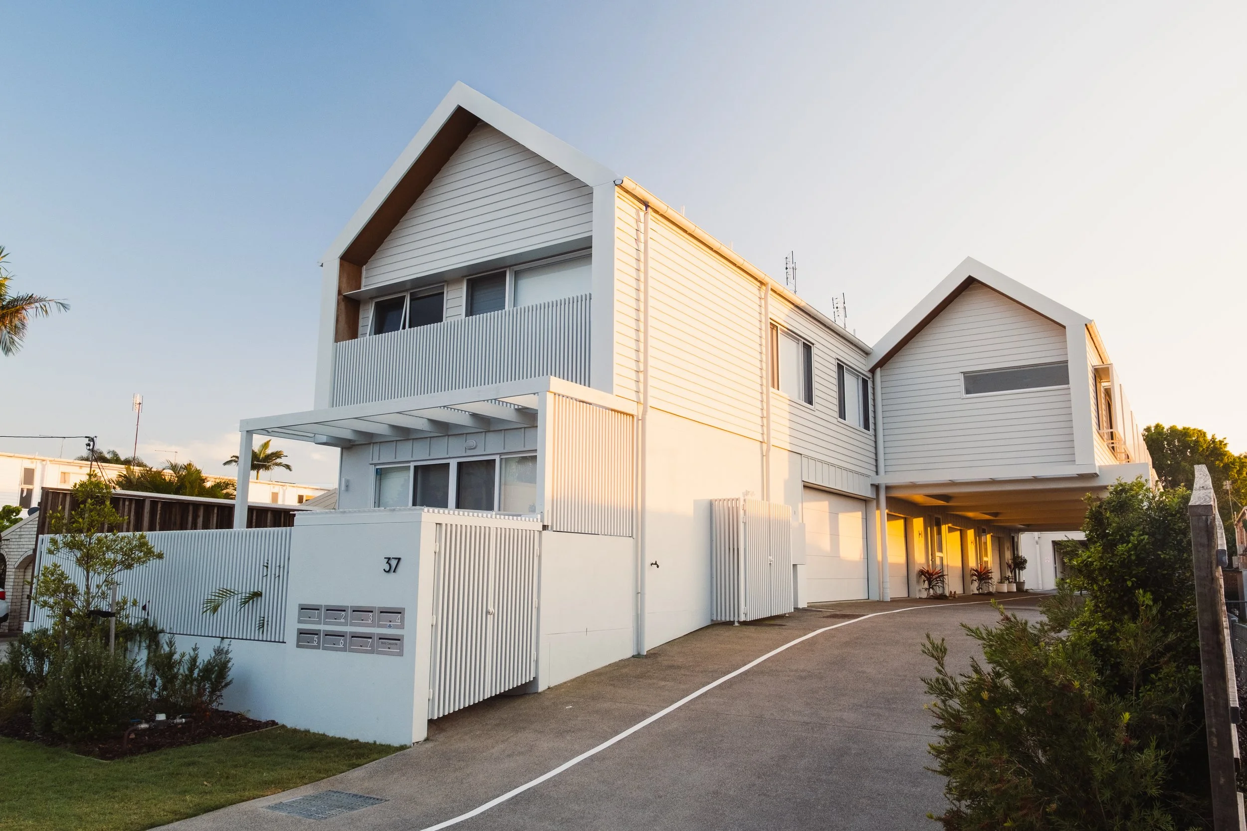 Modern multi-story white residential building with sloped roofs and balconies, situated on a sloped driveway during sunset.