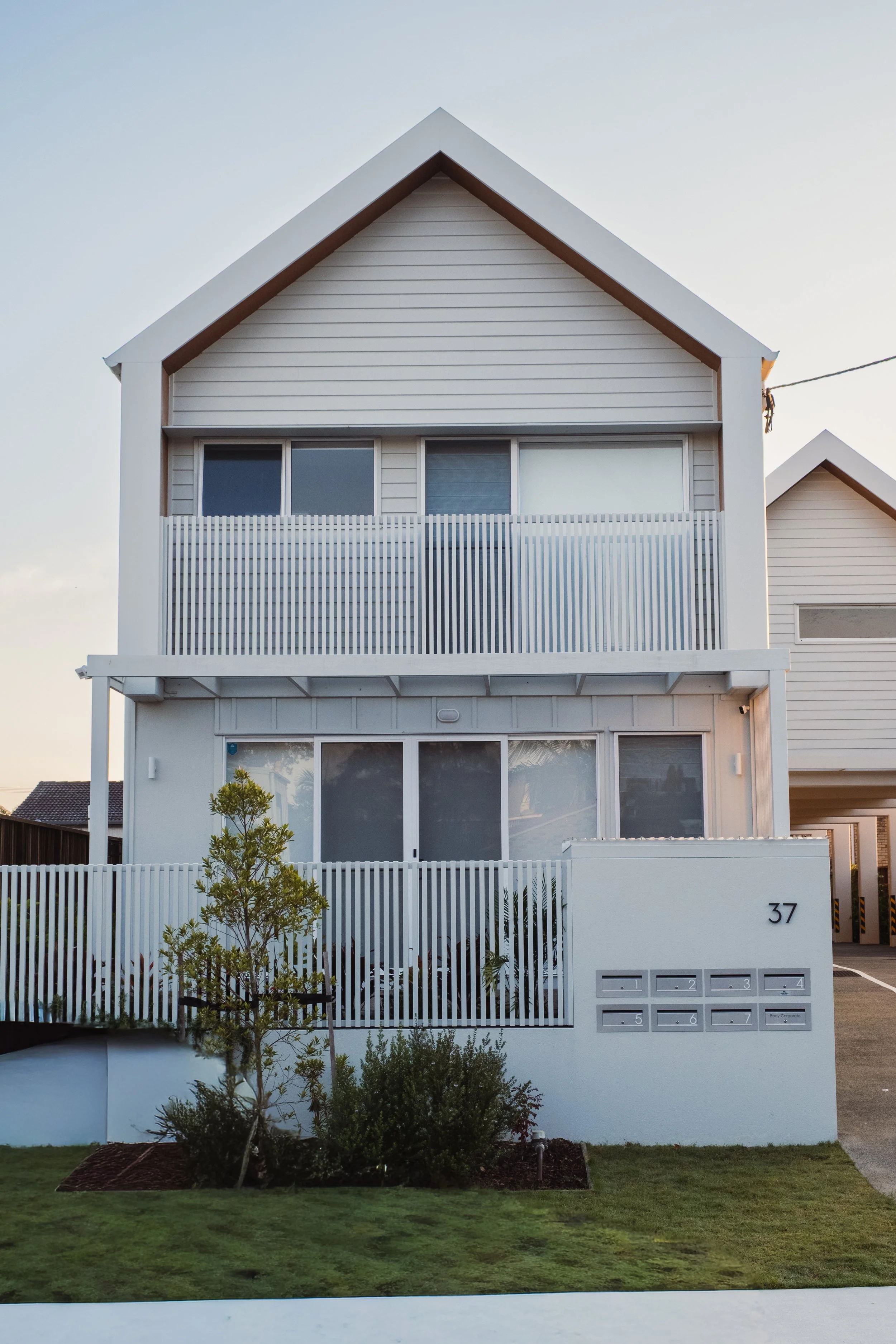 A modern, white, two-story house with a sloped roof and large windows, surrounded by a small yard with bushes and a tree, and a white fence with house number 37 on the wall.
