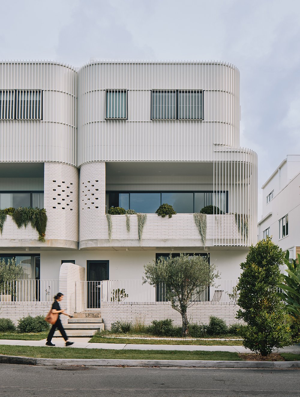 Modern white multi-story building with curved architecture, vertical window blinds, and landscaped balconies. A woman walks past on the sidewalk.