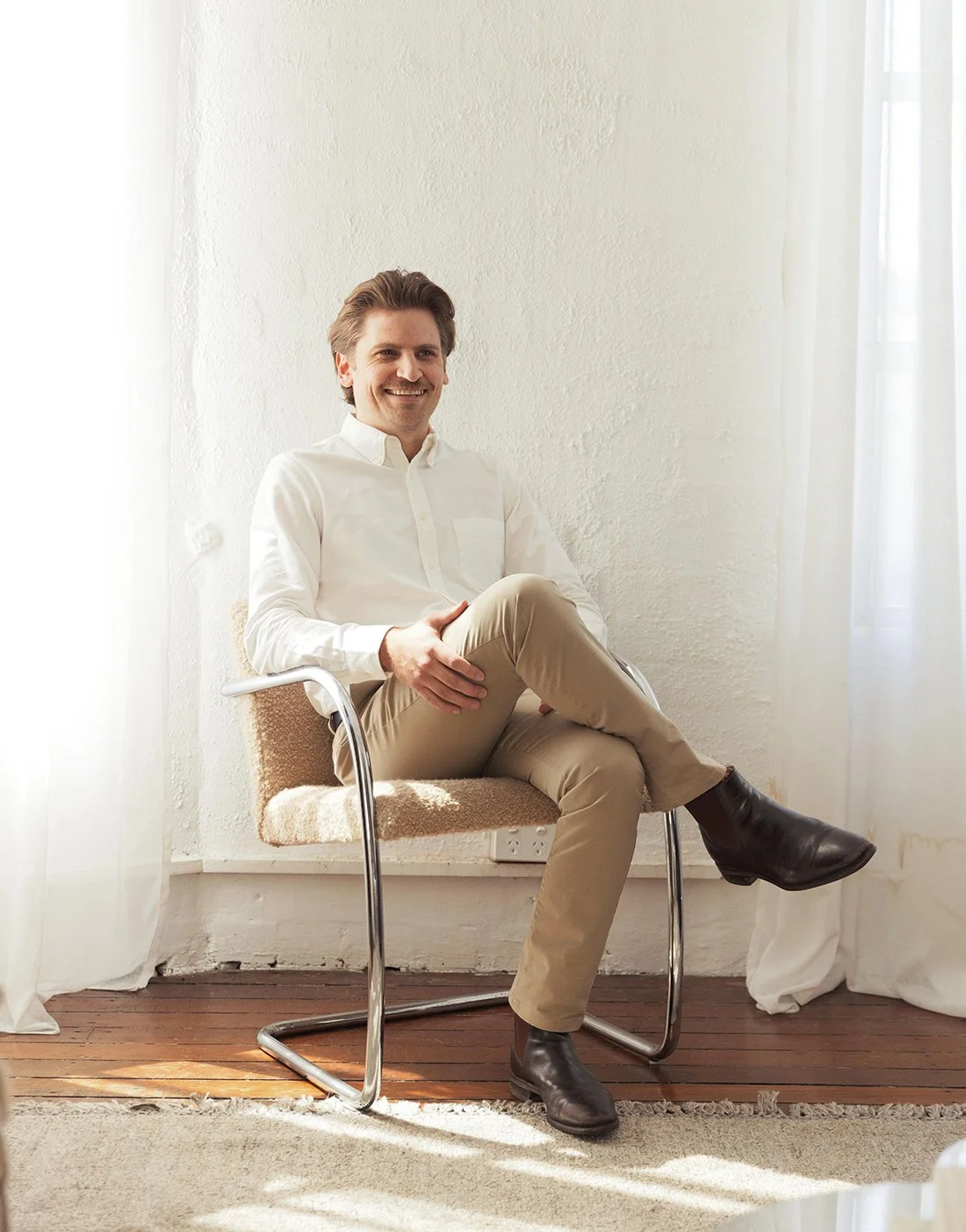 A man sitting on a chair in a bright room with white curtains, wearing a white shirt, beige pants, and dark boots, smiling at the camera.