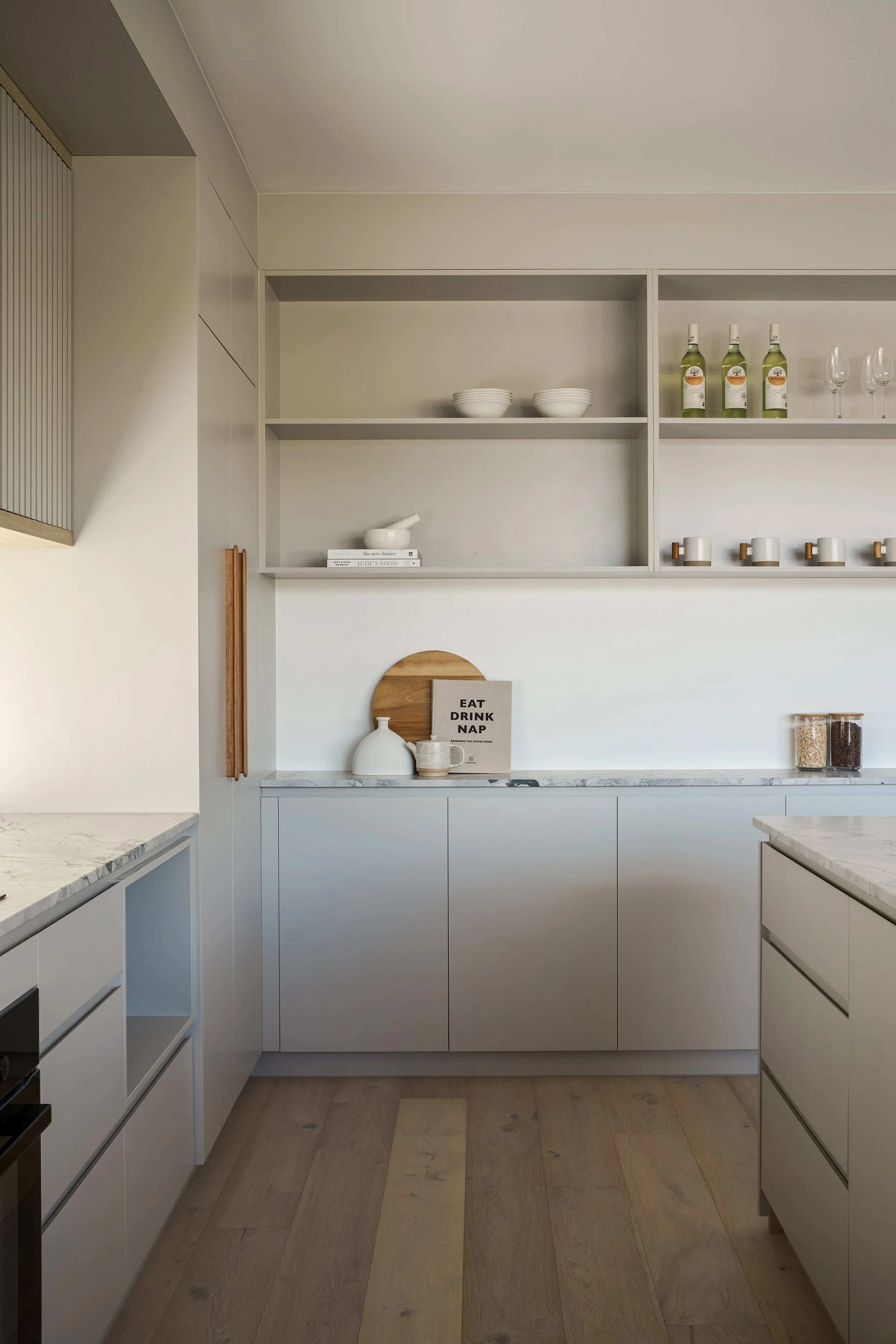 Minimalist kitchen with white cabinets, open shelving, and decorative items including bottles, glasses, bowls, and a sign that says 'EAT DRINK NAP'.