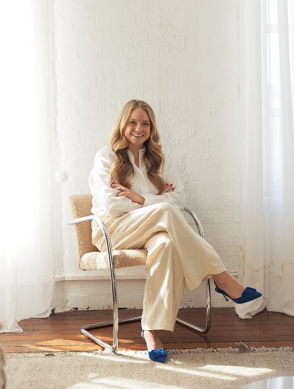A woman with long blonde hair, dressed in a white blouse and cream pants, sitting on a modern beige chair with metal arms, smiling at the camera in bright natural light from nearby window with white curtains.