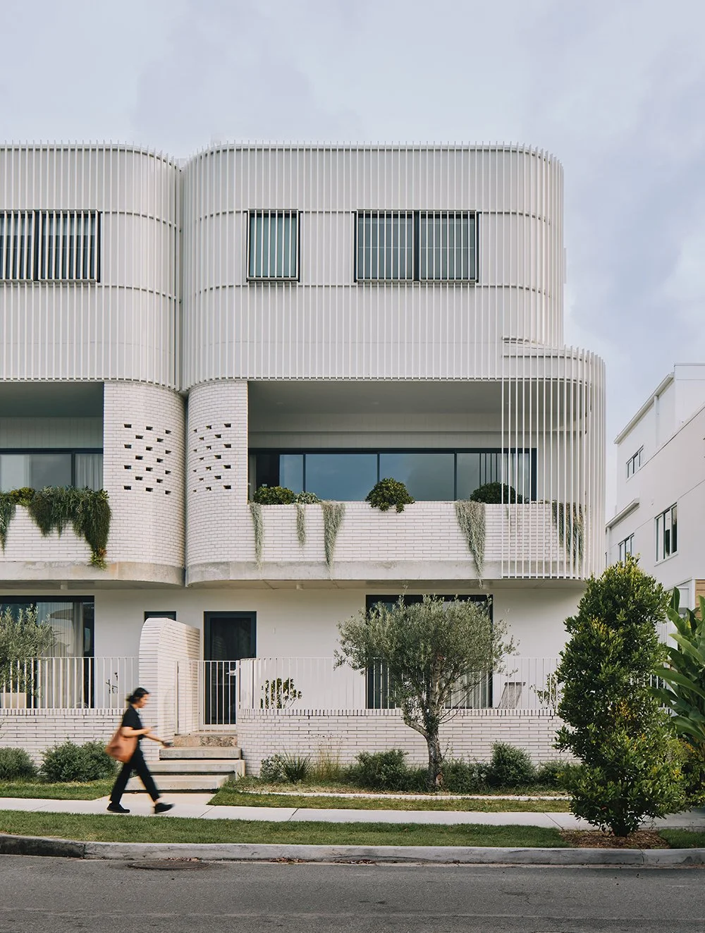Modern white apartment building with curved design, vertical slats, balconies with greenery, and a pedestrian walking by on the sidewalk.