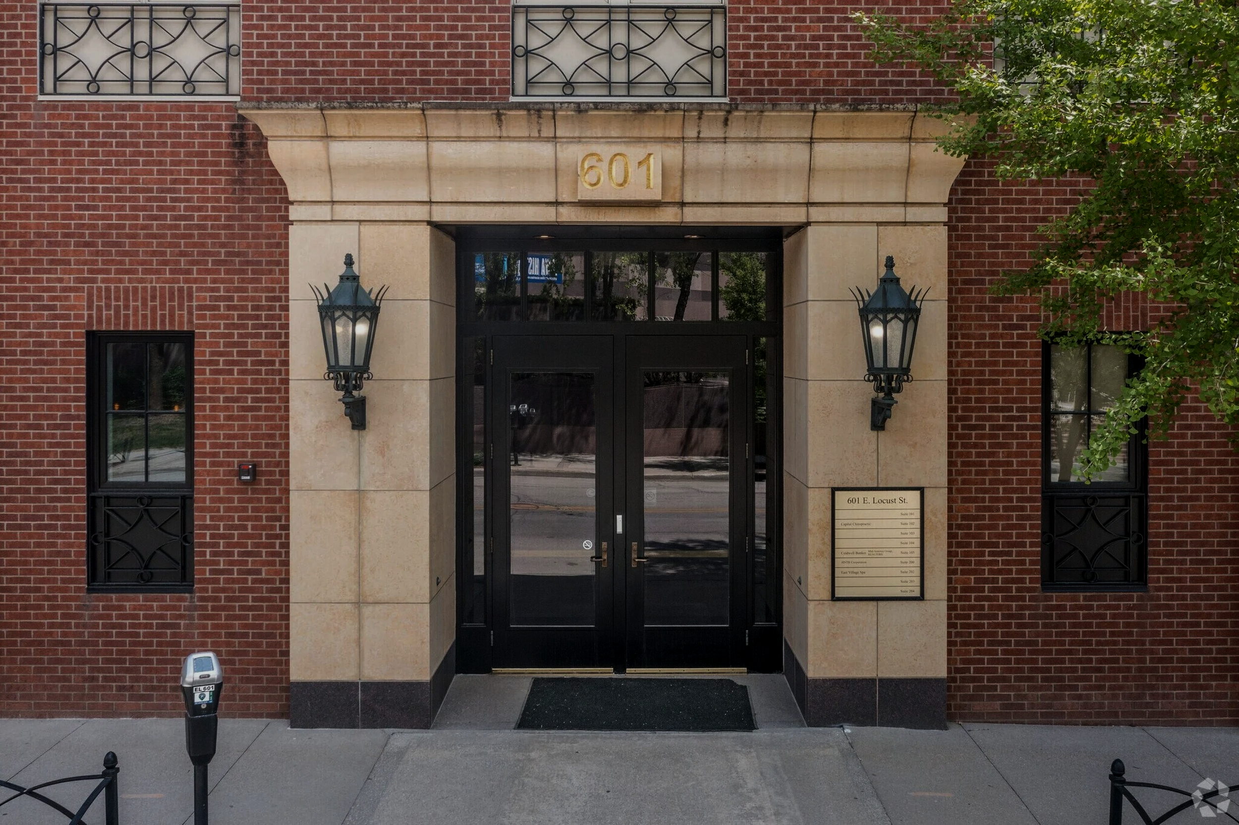 The entrance to a building with the street number 601 displayed above the door. The building has brick walls and black metal window bars. Two wall-mounted lantern-style lights are on either side of the door. There is a tree with green leaves on the right side of the entrance.