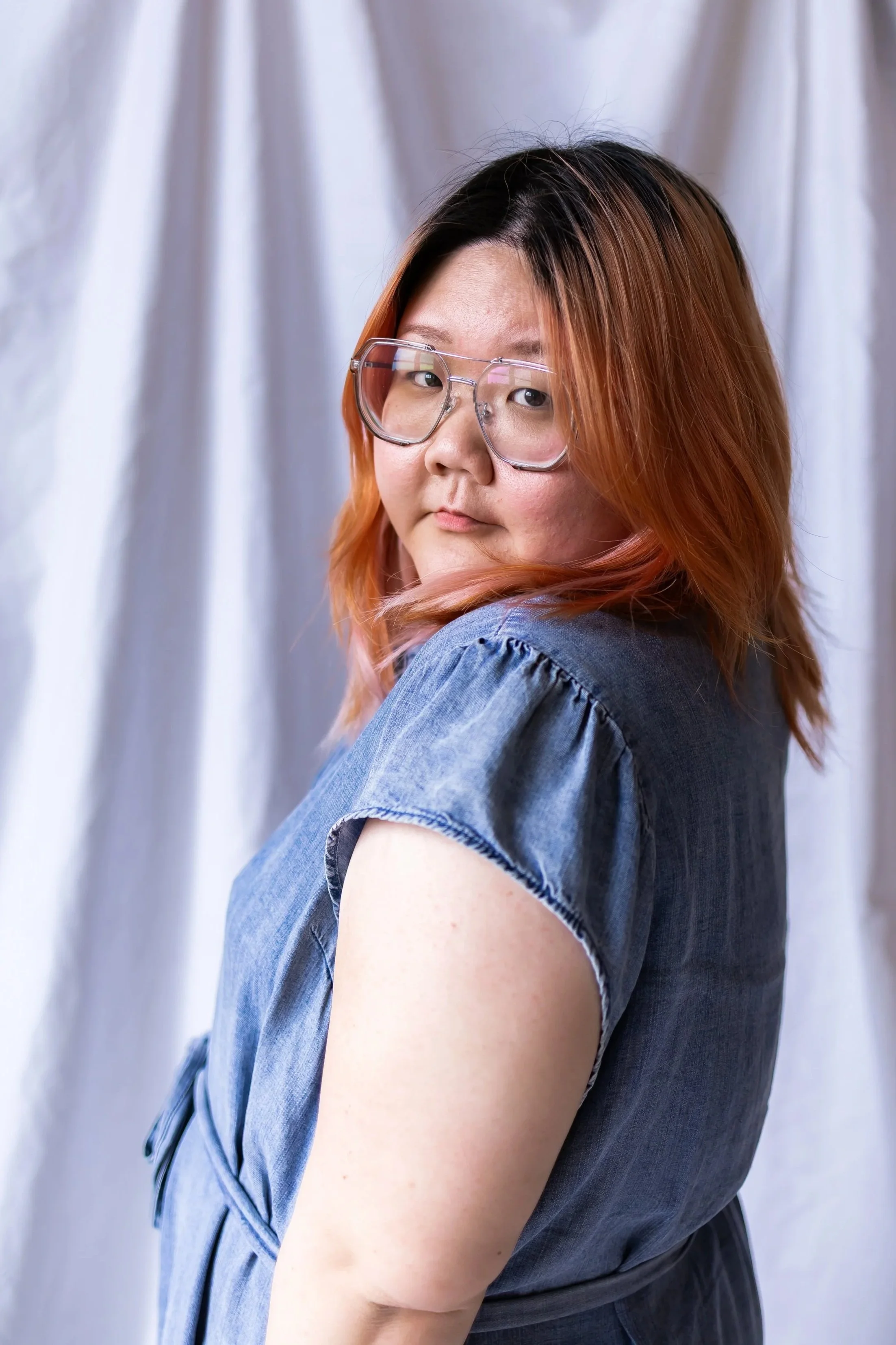 A person with shoulder-length hair, colored with black at the roots and orange at the ends, wearing clear glasses and a denim shirt, standing in front of a light-colored curtain.