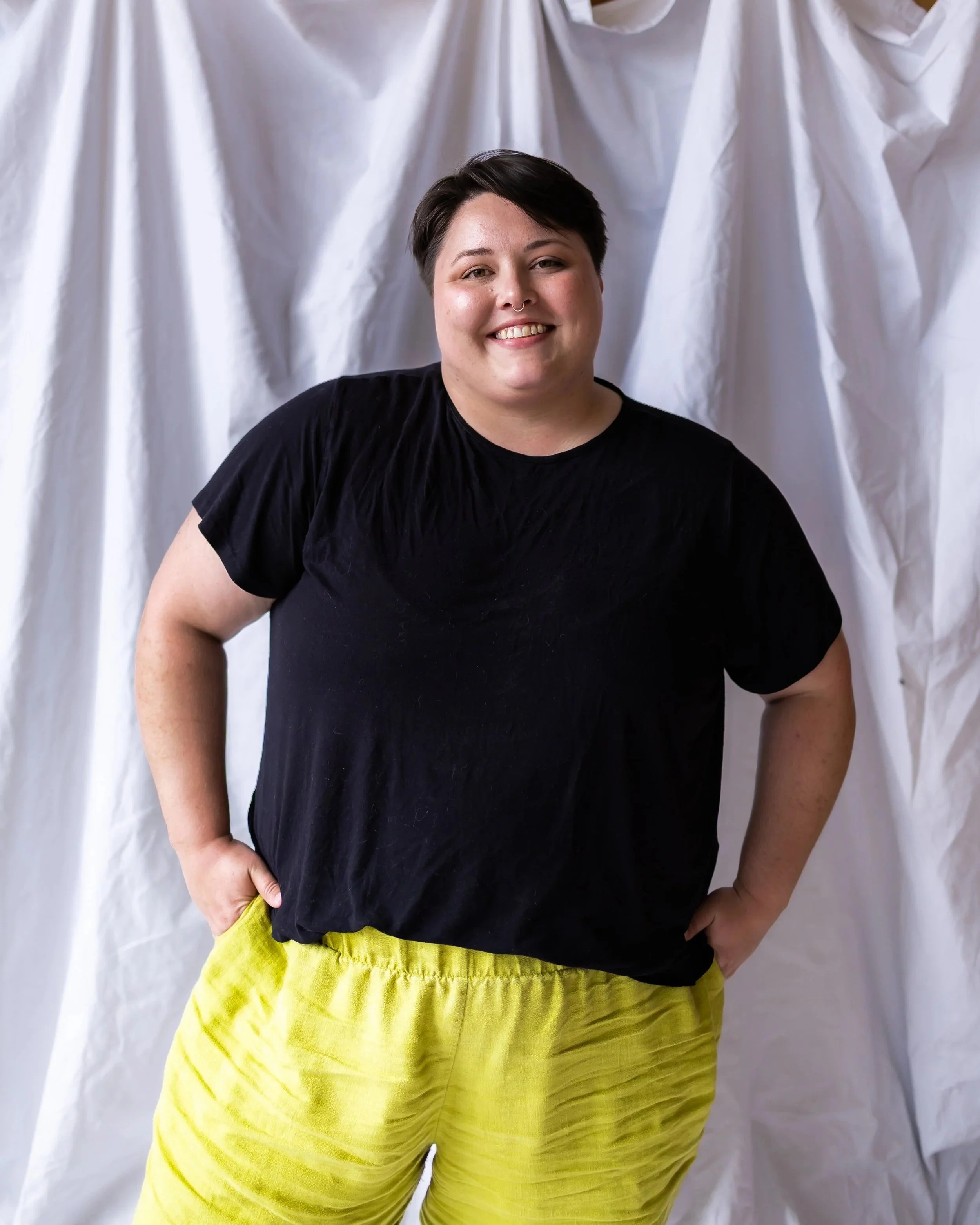 Person with short dark hair smiling, wearing a black T-shirt and bright yellow shorts, standing in front of a white fabric backdrop.