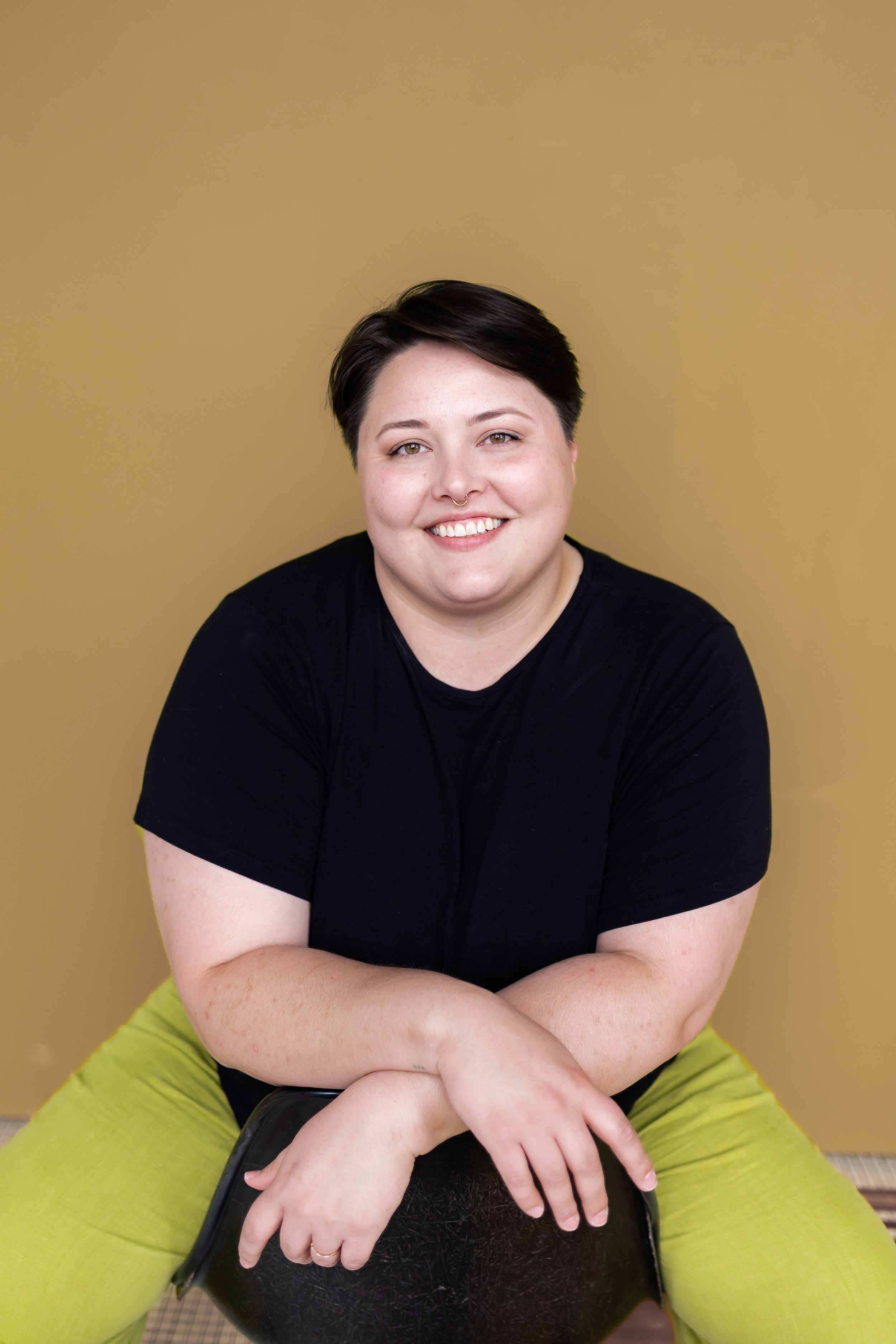 A person with short dark hair, a septum piercing, and light skin, smiling with makeup, wearing a black t-shirt and green pants, sitting cross-legged on a chair against a beige background.