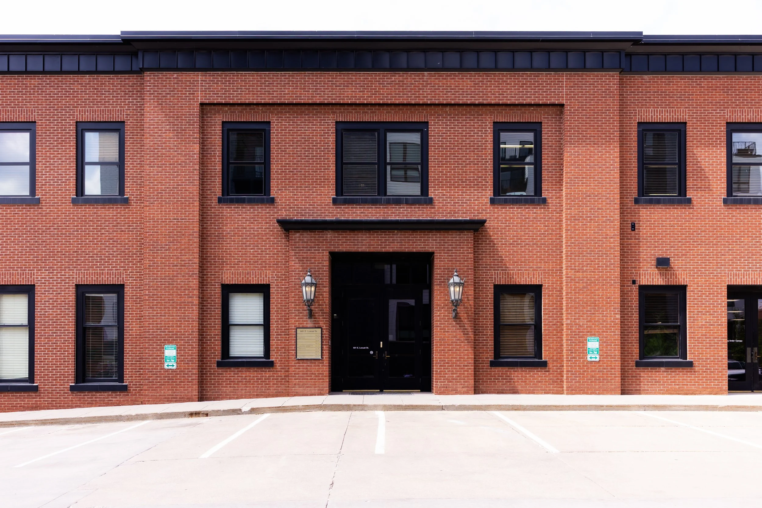 Red brick building with black-framed windows and black doors, two lamps outside, and parking lot in front.