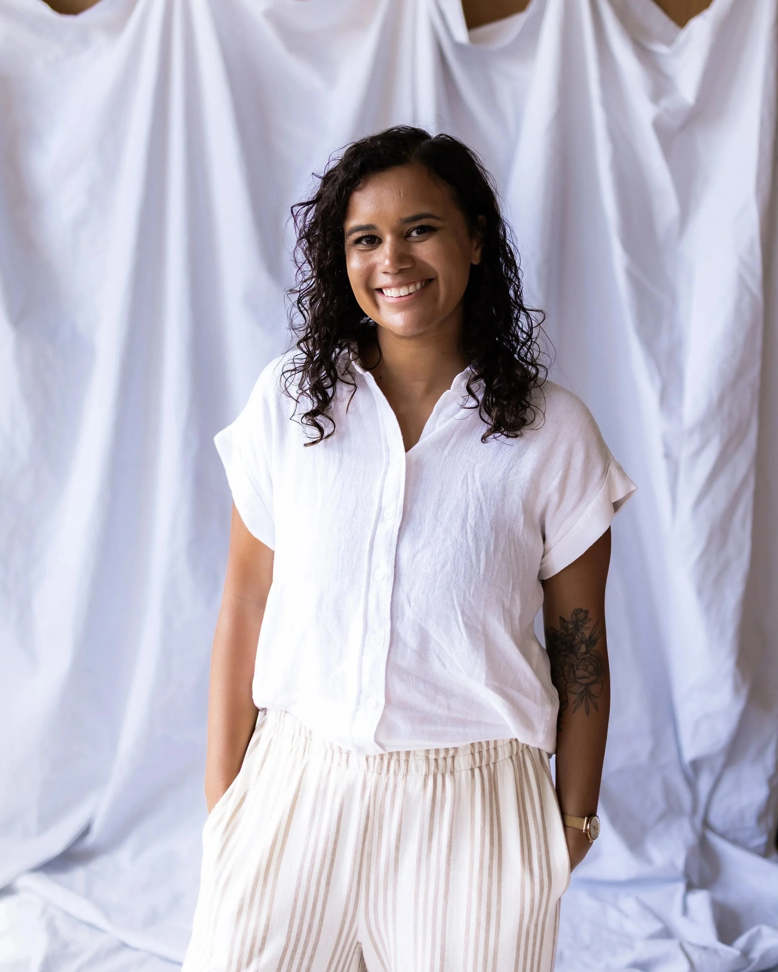 A woman with dark curly hair, wearing a white button-up shirt with rolled sleeves, beige and white striped pants, a gold watch, and a tattoo on her left forearm. She is standing against a white crumpled fabric backdrop and smiling.