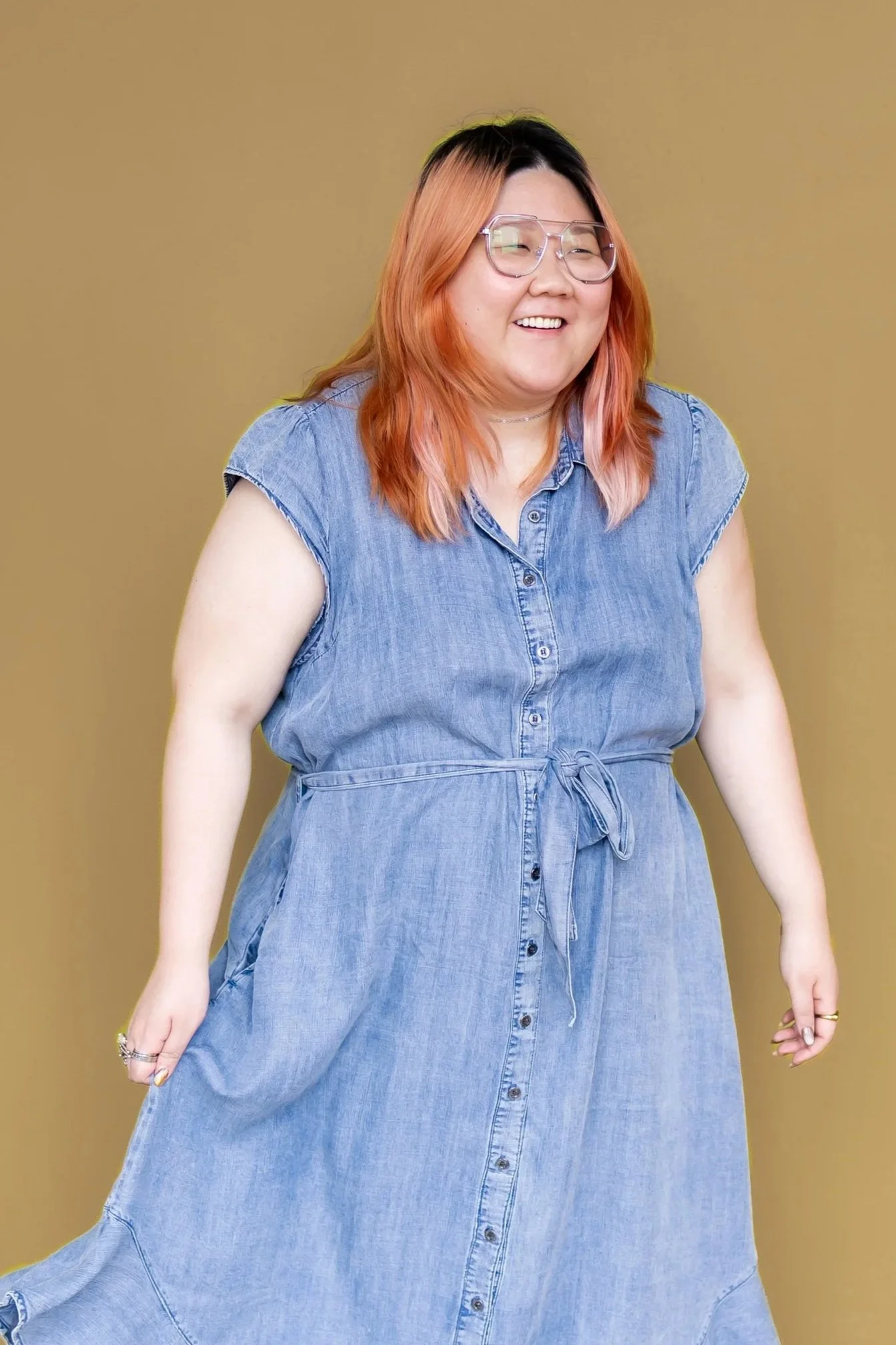 Smiling woman with pink hair, glasses, wearing a denim dress, standing against a tan background.