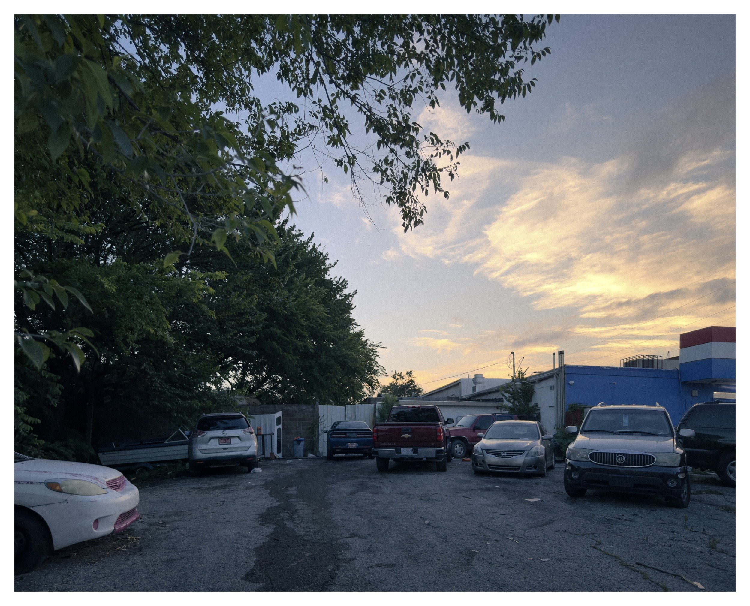 An outdoor parking lot filled with cars during the evening, with trees and a building in the background under a sky with scattered clouds.