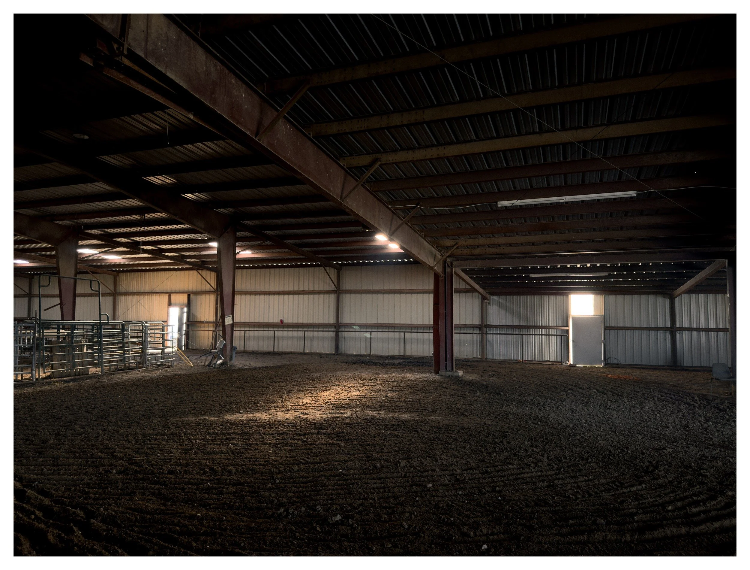 Interior view of an empty indoor horse riding arena or barn with a dirt floor, metal beams, and a corrugated metal ceiling.