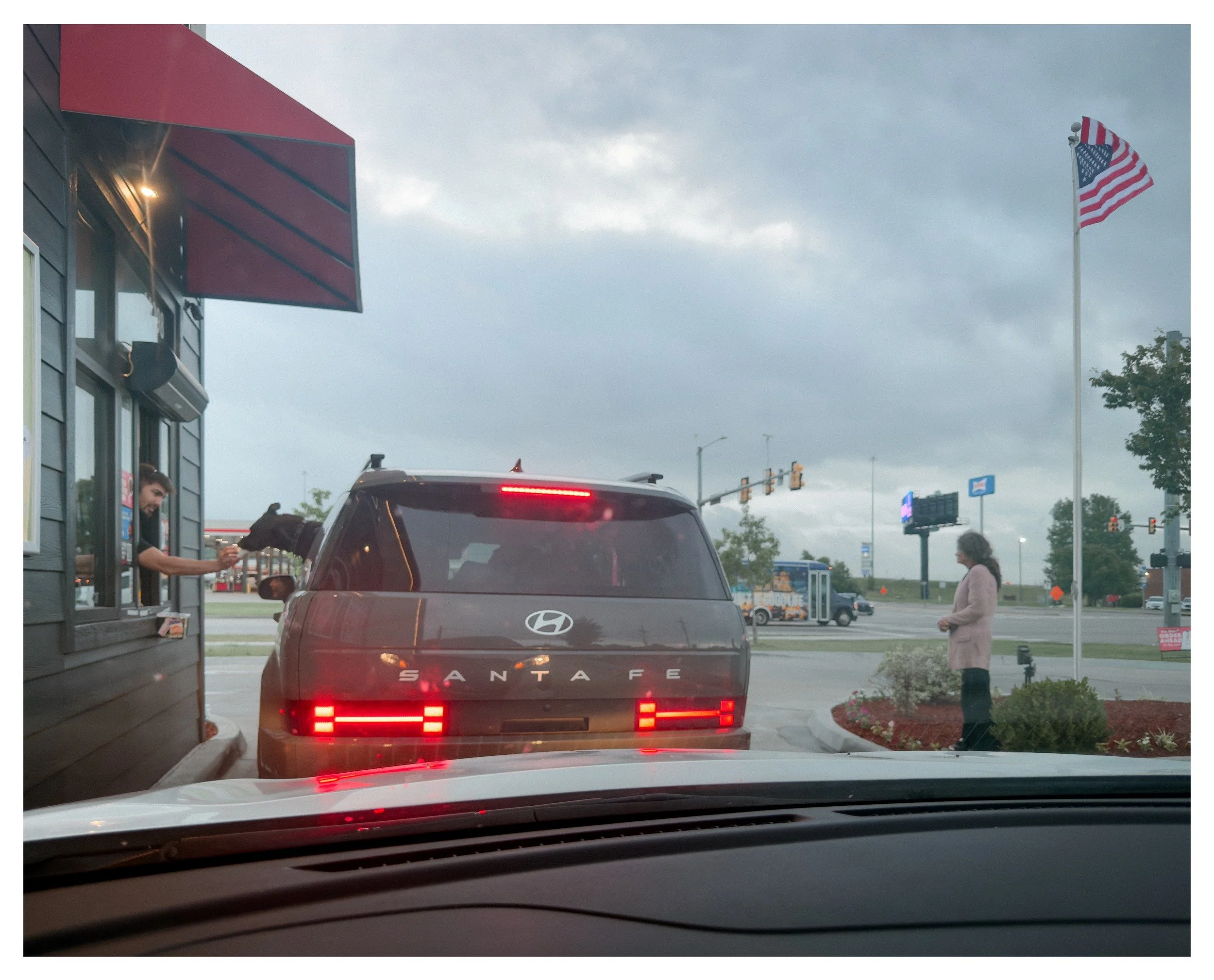 The image shows a view from inside a car waiting in line at a drive-thru, with a black Hyundai Santa Fe in front. There is a fast food restaurant on the left side with a red awning and a person taking an order. A flagpole with the American flag is on