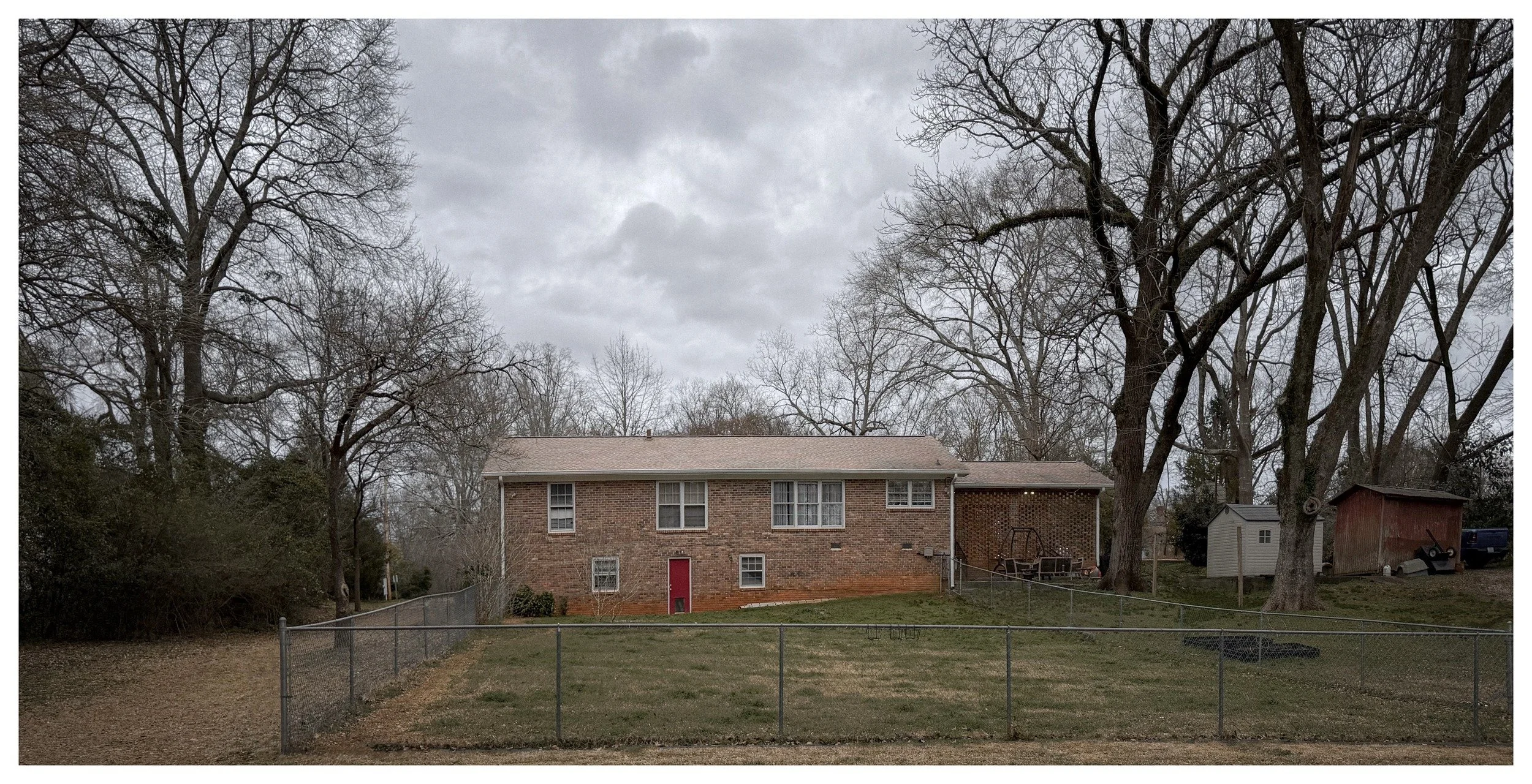 A brick house with a red door, surrounded by a fenced backyard with grass and large leafless trees, under a cloudy sky.