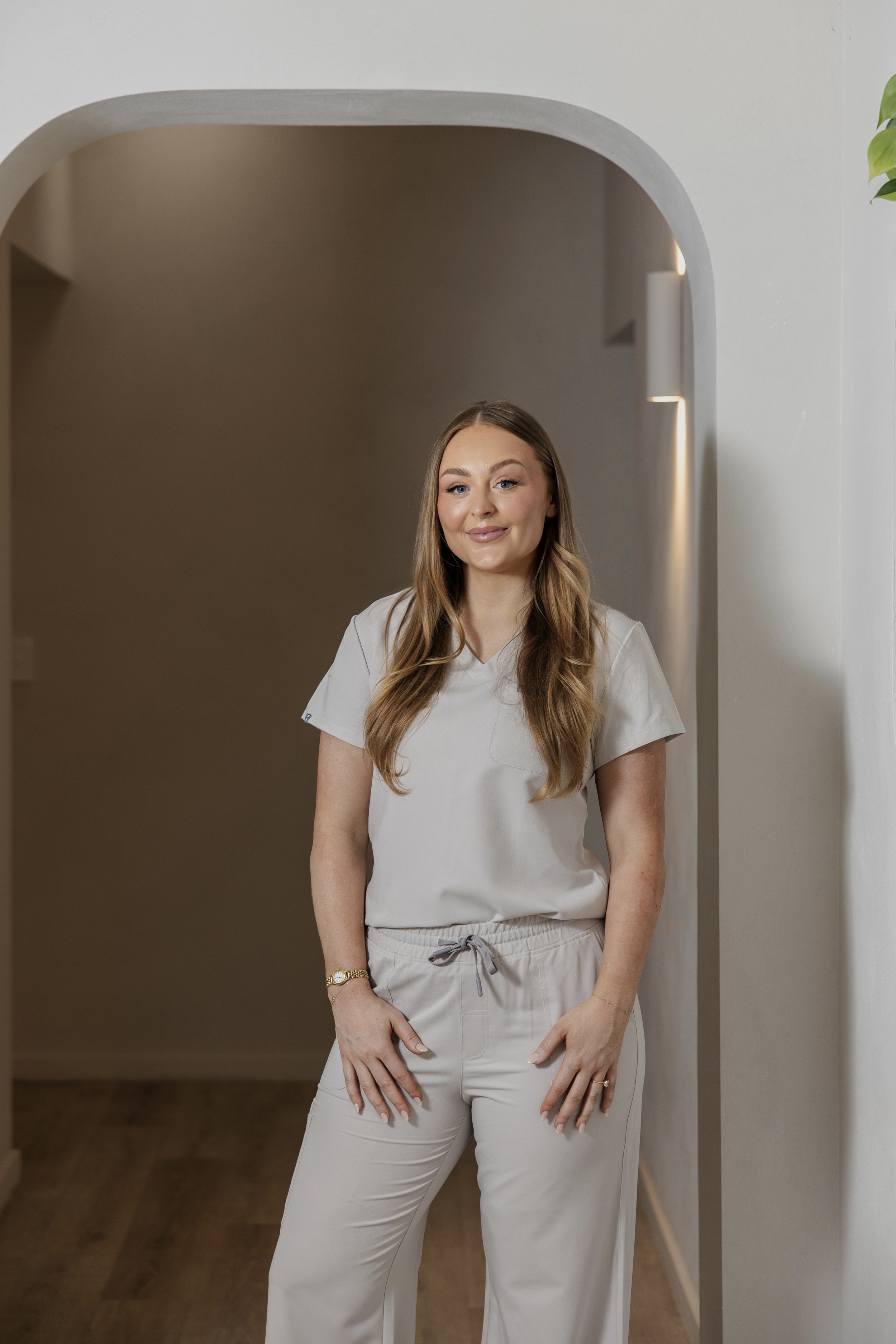 A young woman in beige scrubs standing in a hallway with warm lighting and an arched doorway.