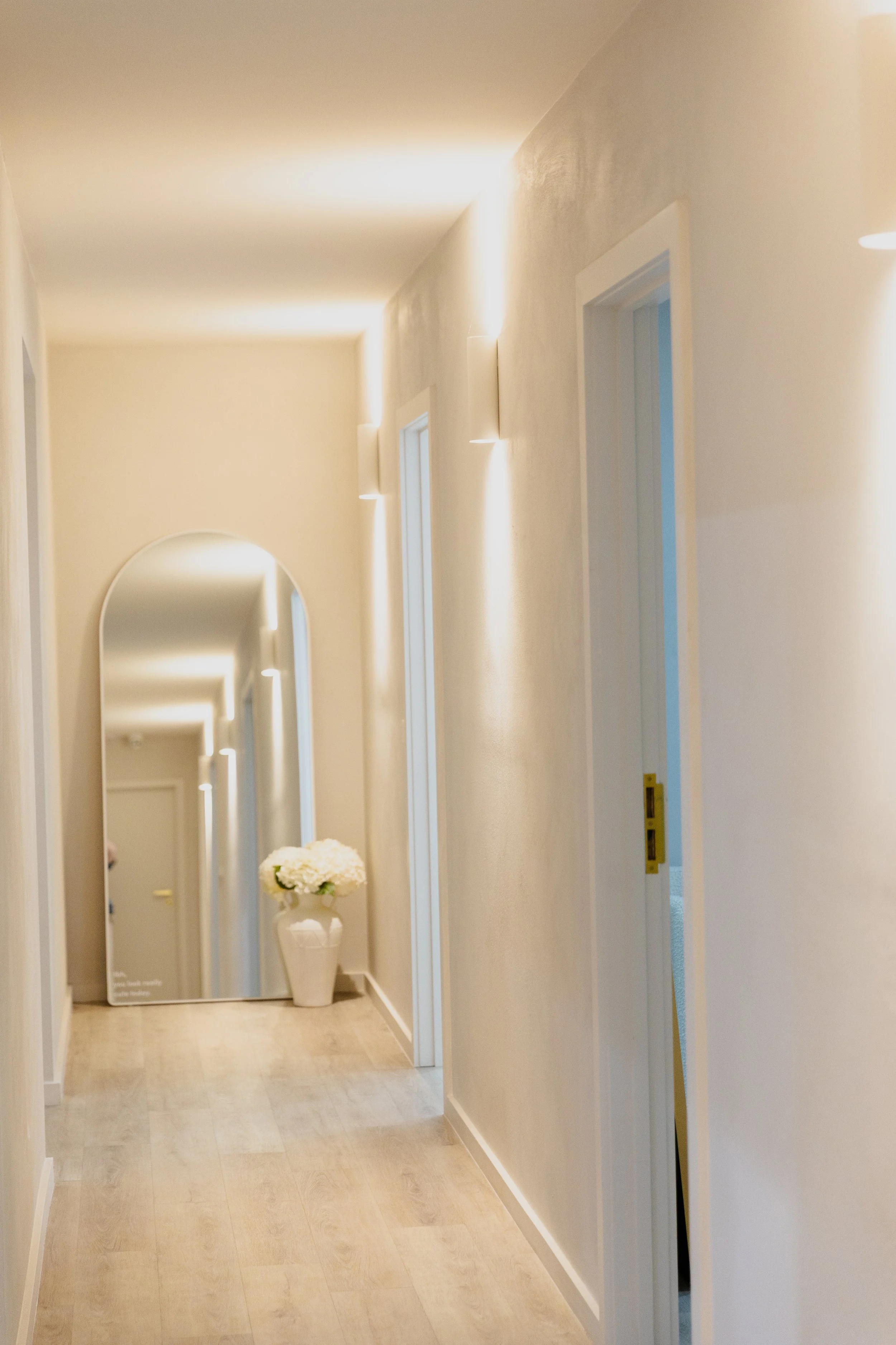A hallway with light-colored walls and wooden flooring, featuring wall-mounted lights, and a large arched mirror at the end of the corridor with a white flower arrangement in a vase beside it.