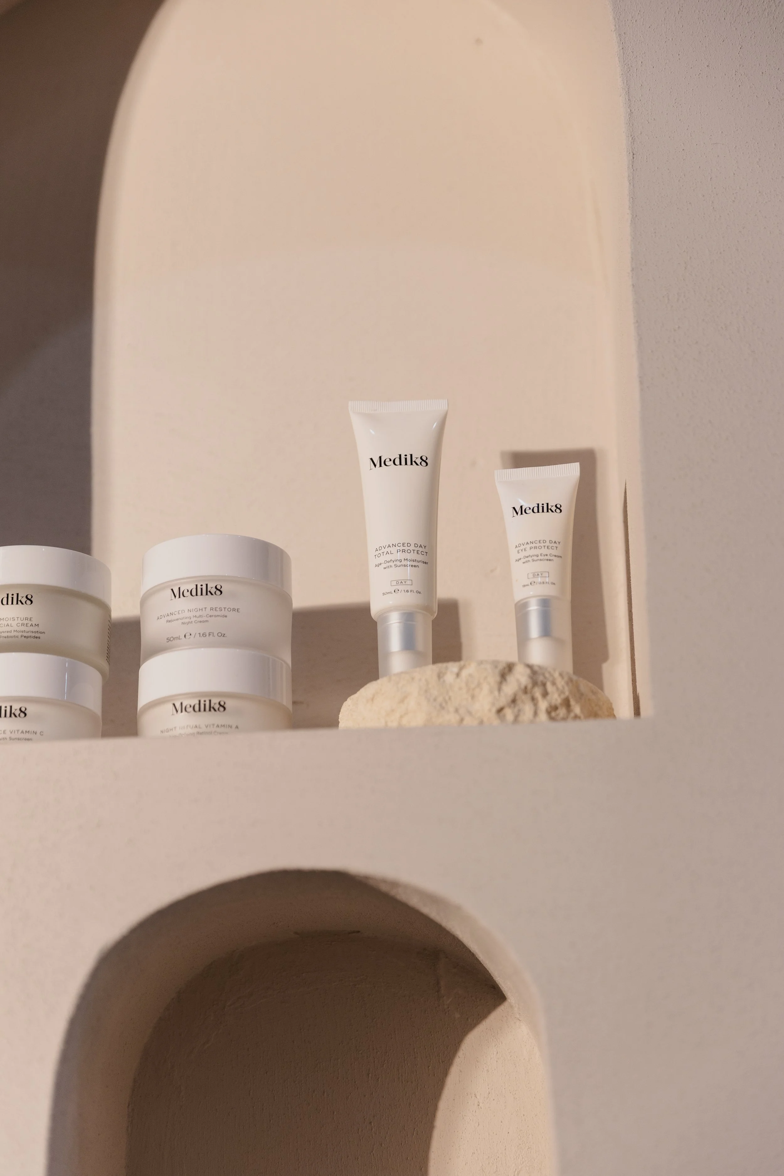 Collection of Medik8 skincare products arranged on a beige shelf with a decorative stone, including creams and sunscreens.