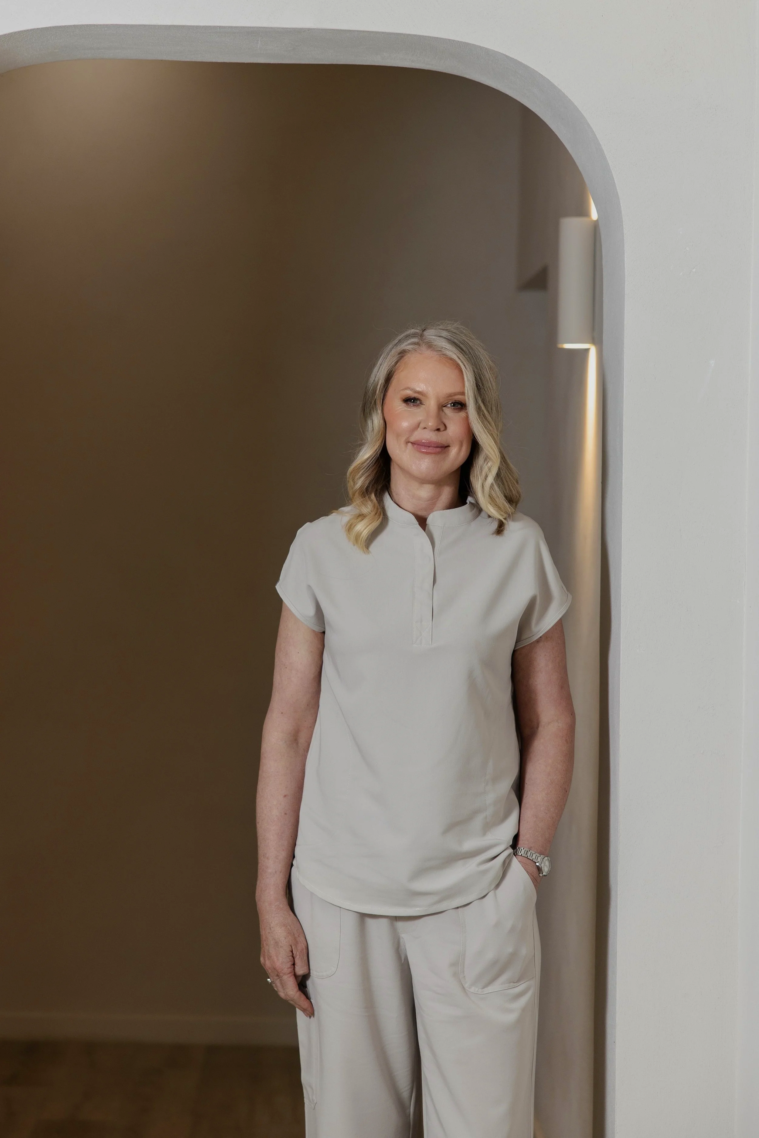 A woman with blonde hair wearing beige scrubs standing in a well-lit modern room with white walls and wooden flooring.