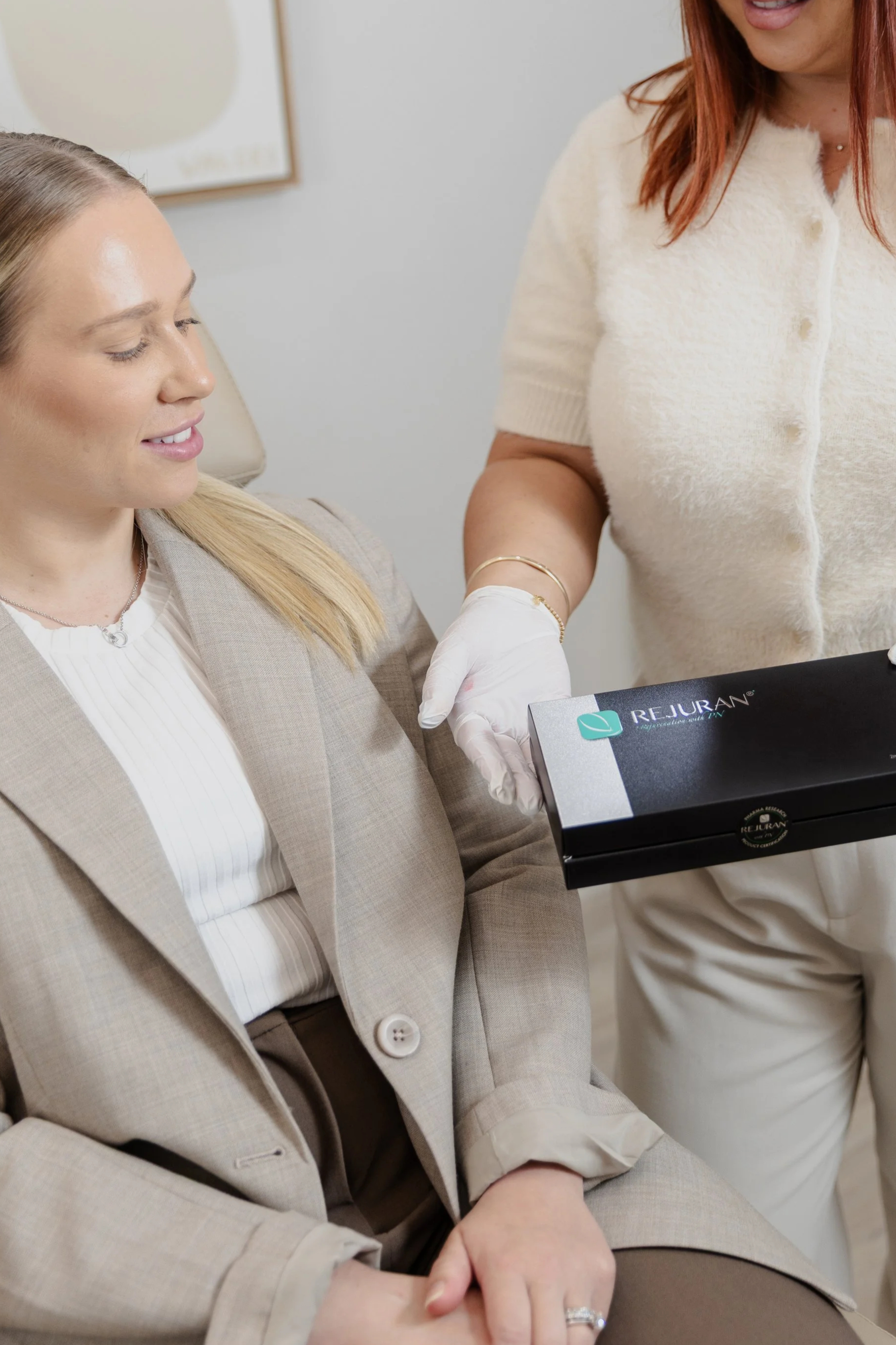 A woman in a beige blazer sitting on a chair, receiving a cosmetic treatment in her hand from a healthcare professional wearing white gloves. The professional is holding a black box labeled 'Rejuran'.