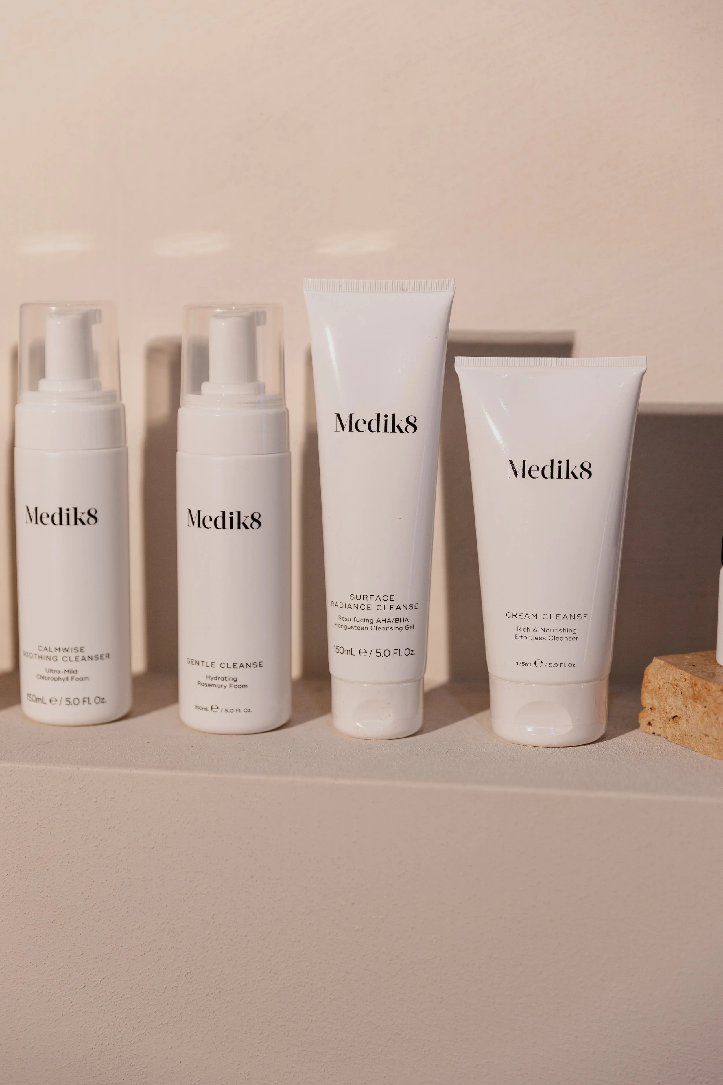 A row of four white skincare product bottles and tubes from Medik8 on a beige shelf, with soft lighting and a neutral background.