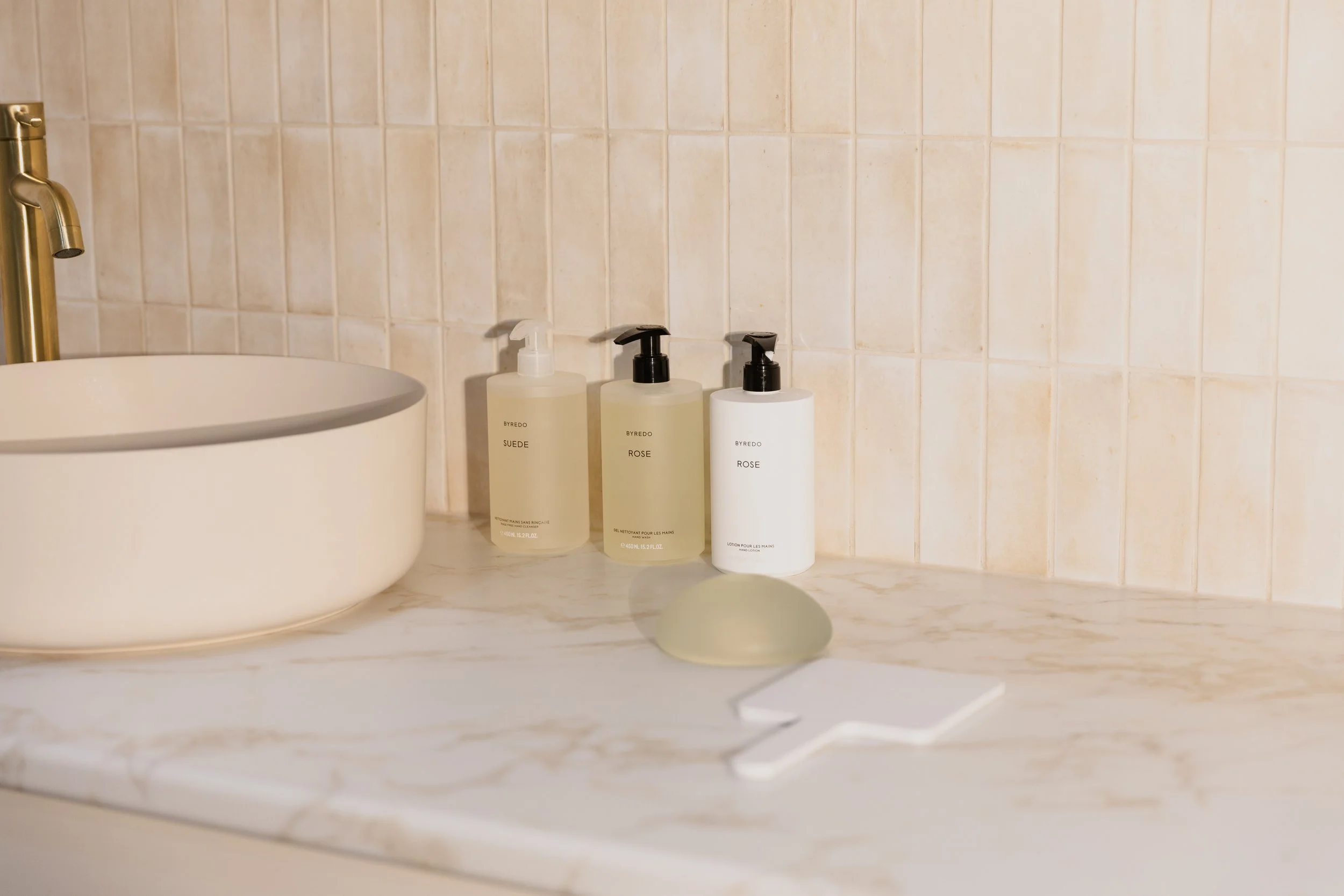 Bathroom countertop with three soap bottles, a soap dish, and a white sink with a gold faucet, beige tiled wall background.