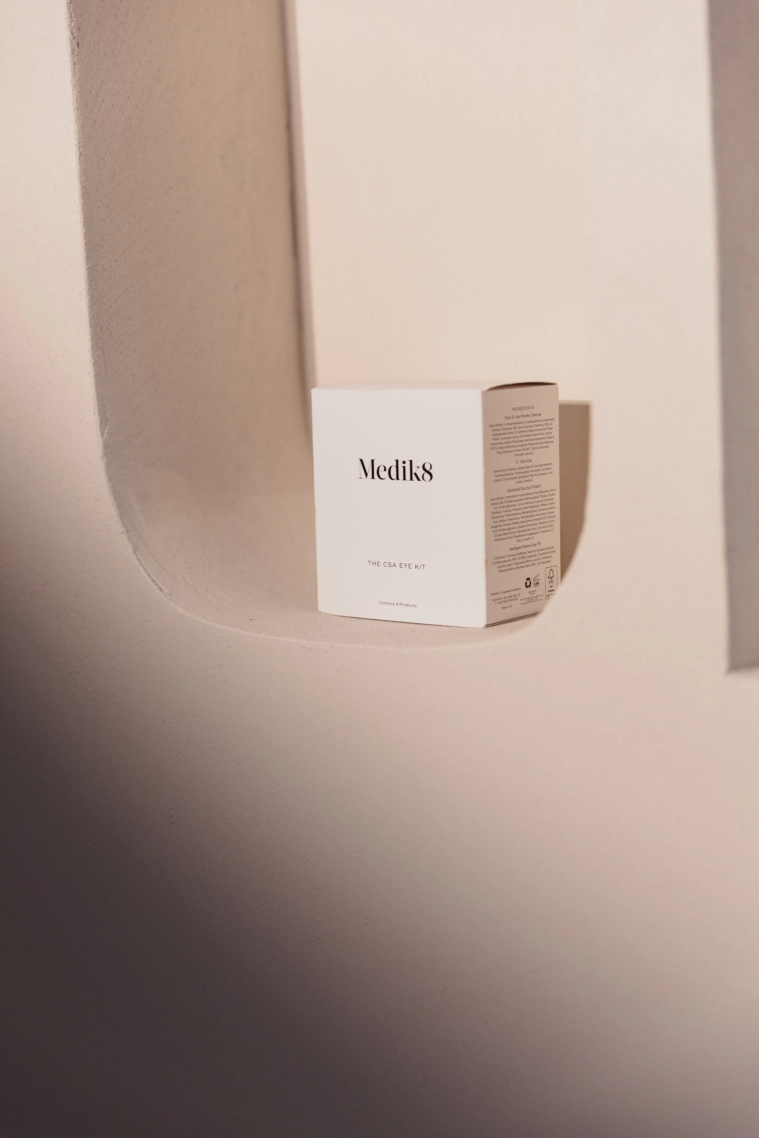 A white box labeled 'Medik8' with additional text about an eye kit, placed on a wooden shelf with a curved edge, in a neutral-colored room.