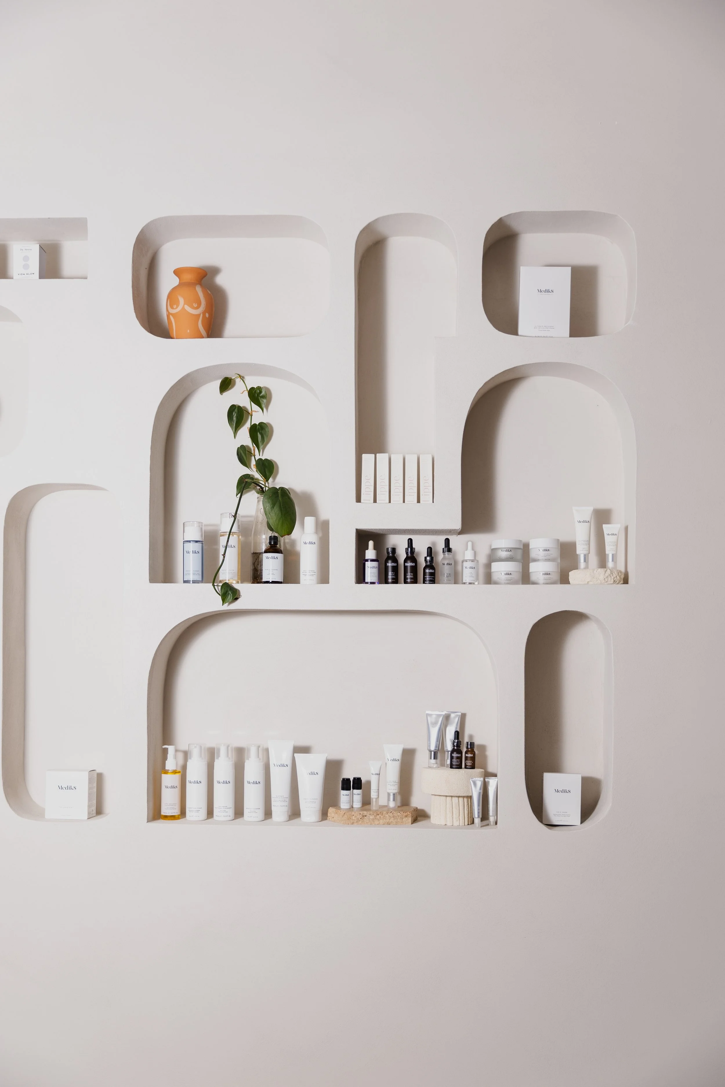 Minimalist display of skincare and beauty products on white built-in wall shelves with a small potted plant and a decorative vase.