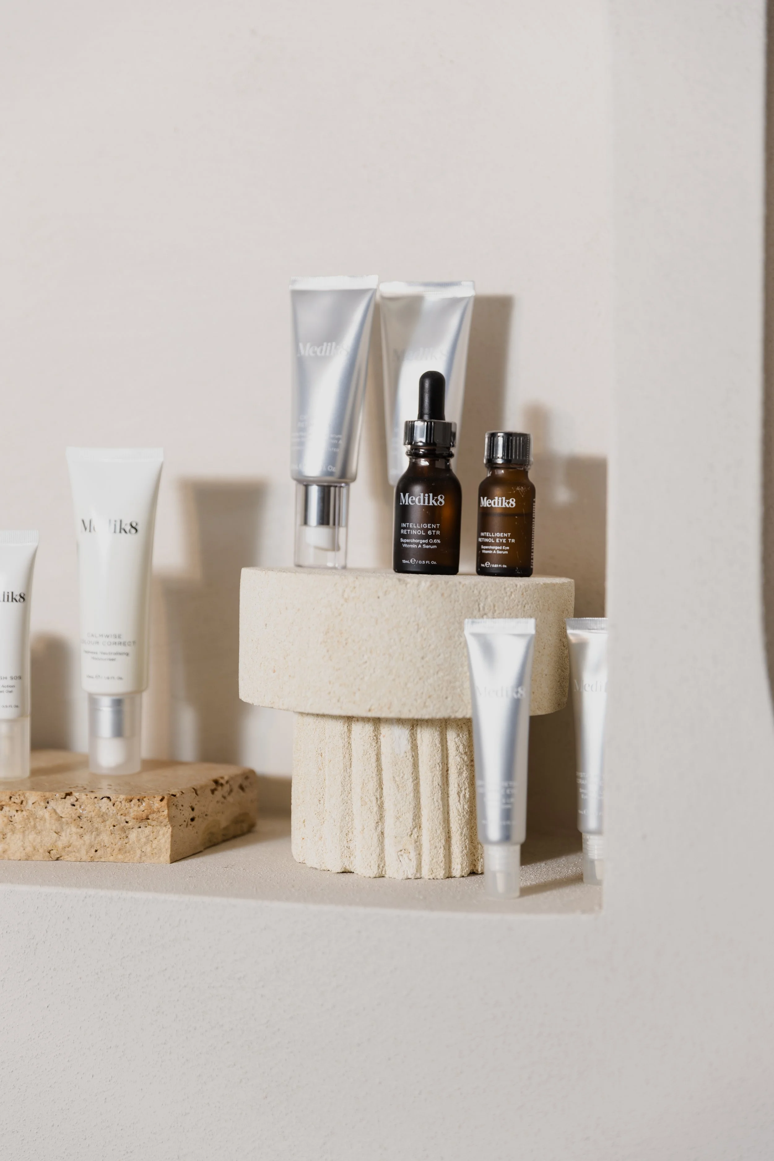 Skincare products displayed on minimalist stands, including tubes, bottles, and dropper bottles, in neutral tones.