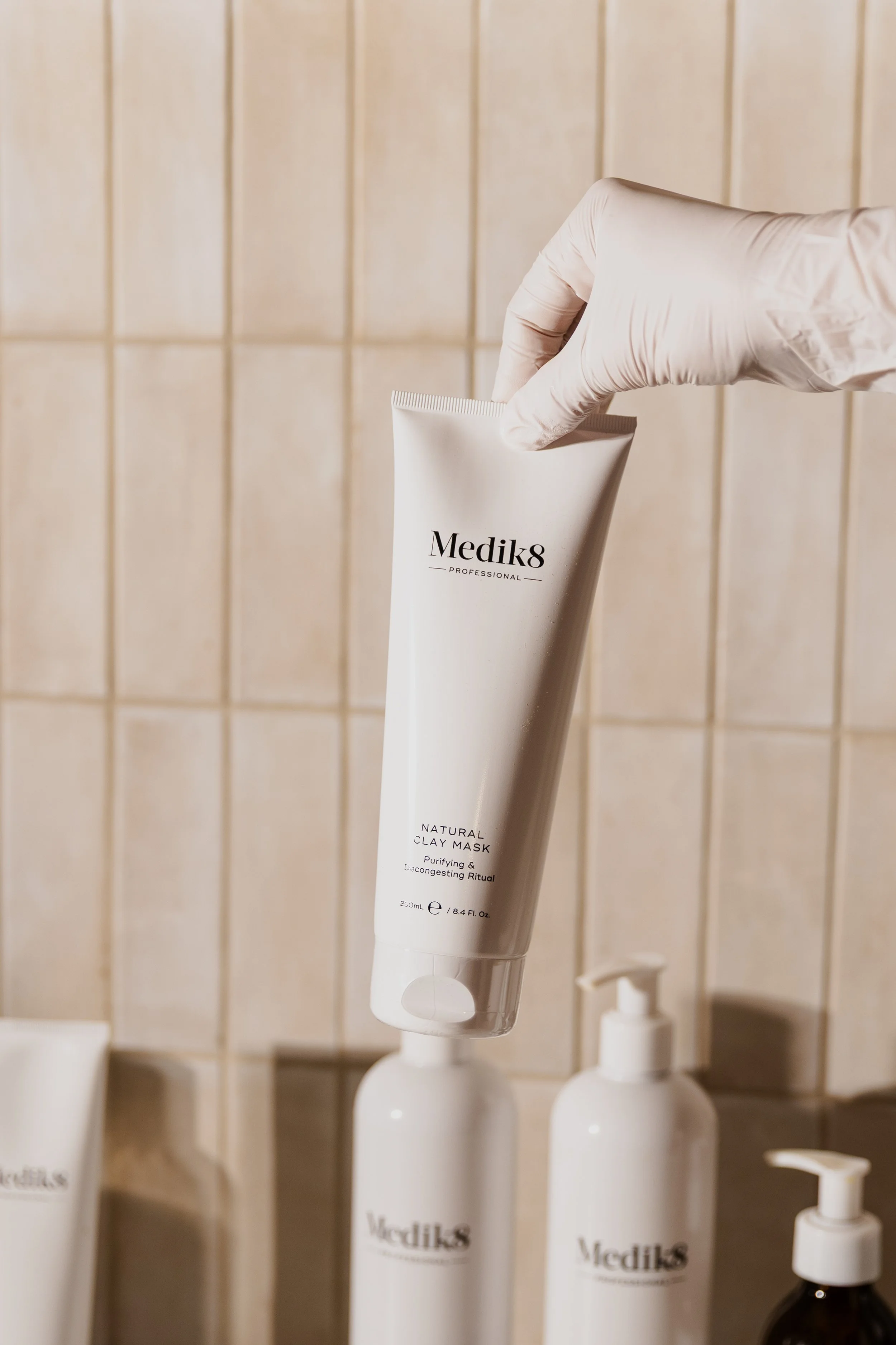 A person wearing a white glove is holding a tube of Medik8 Natural Clay Mask in front of beige tiled wall, with other skincare bottles on a shelf below.